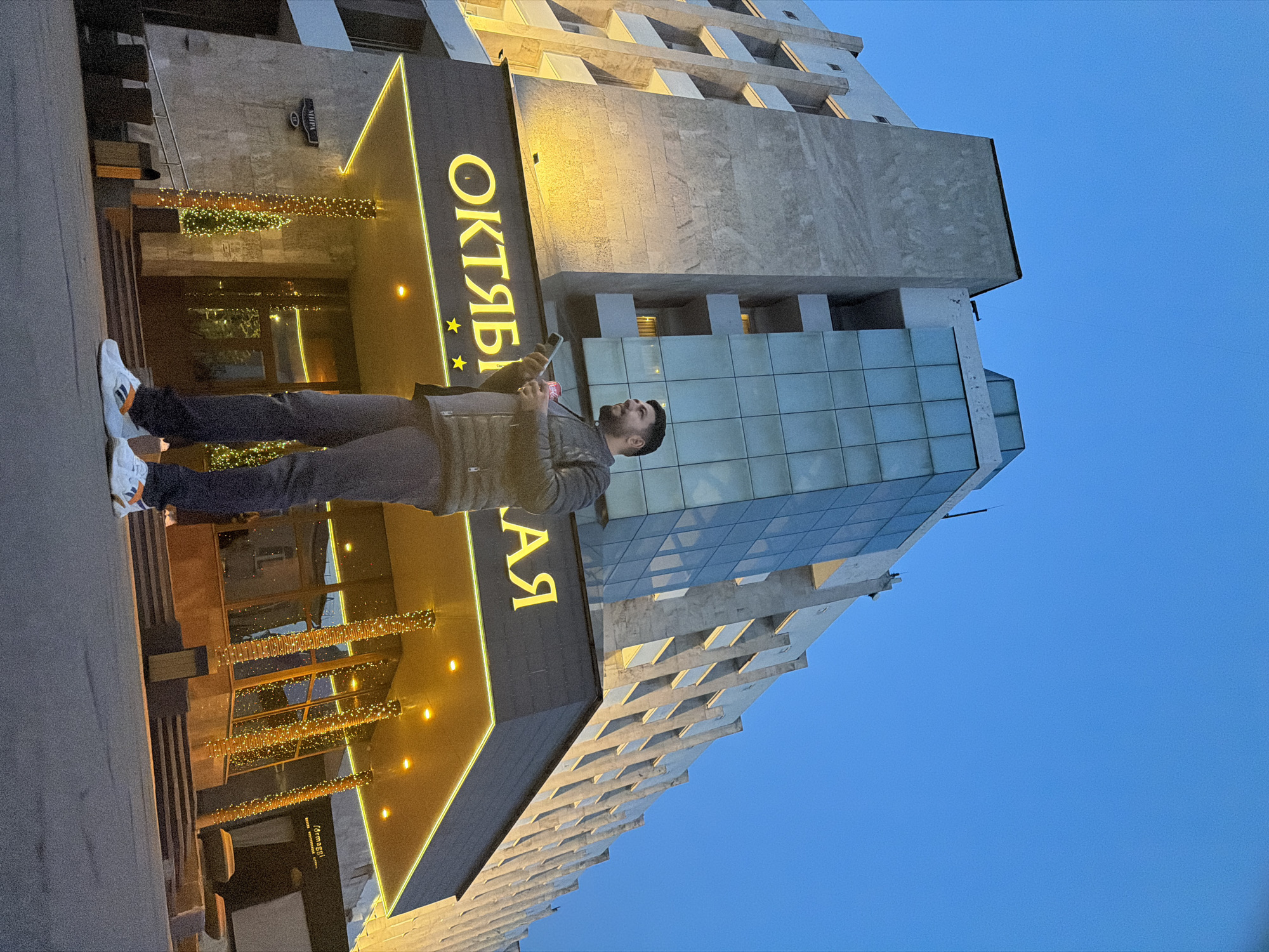 A person stands in front of the Oktyabrskaya Hotel in Krasnoyarsk, Russia. The hotel's sign is prominently displayed. The sky is a clear blue. The person is holding a phone.