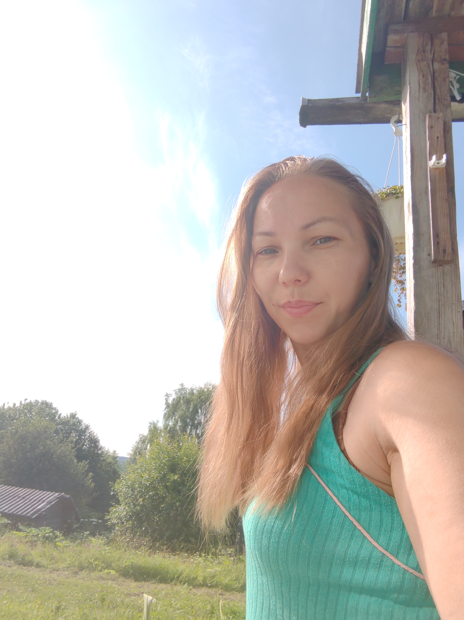 A woman with long, light brown hair is taking a selfie outdoors. She is wearing a teal tank top. Behind her, a wooden structure is visible, with a bright sky and clouds above. The background shows a rural landscape with trees and a small building. The image appears to be taken on a sunny day. The location is specified as Chuchlema, Russia.