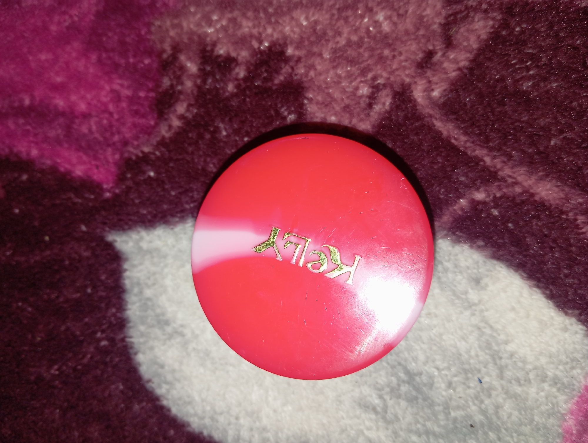 A red, round object, possibly a container lid with the gold lettering "KELLY" embossed on it, rests on a textured surface that appears to be a rug or blanket with shades of purple and white. The object is positioned in the upper left quadrant of the image. The lighting suggests it may be an indoor setting. There are no people or other distinguishing features in the image. The location of Rejosari, Indonesia, cannot be determined from the visual information provided in the image.
