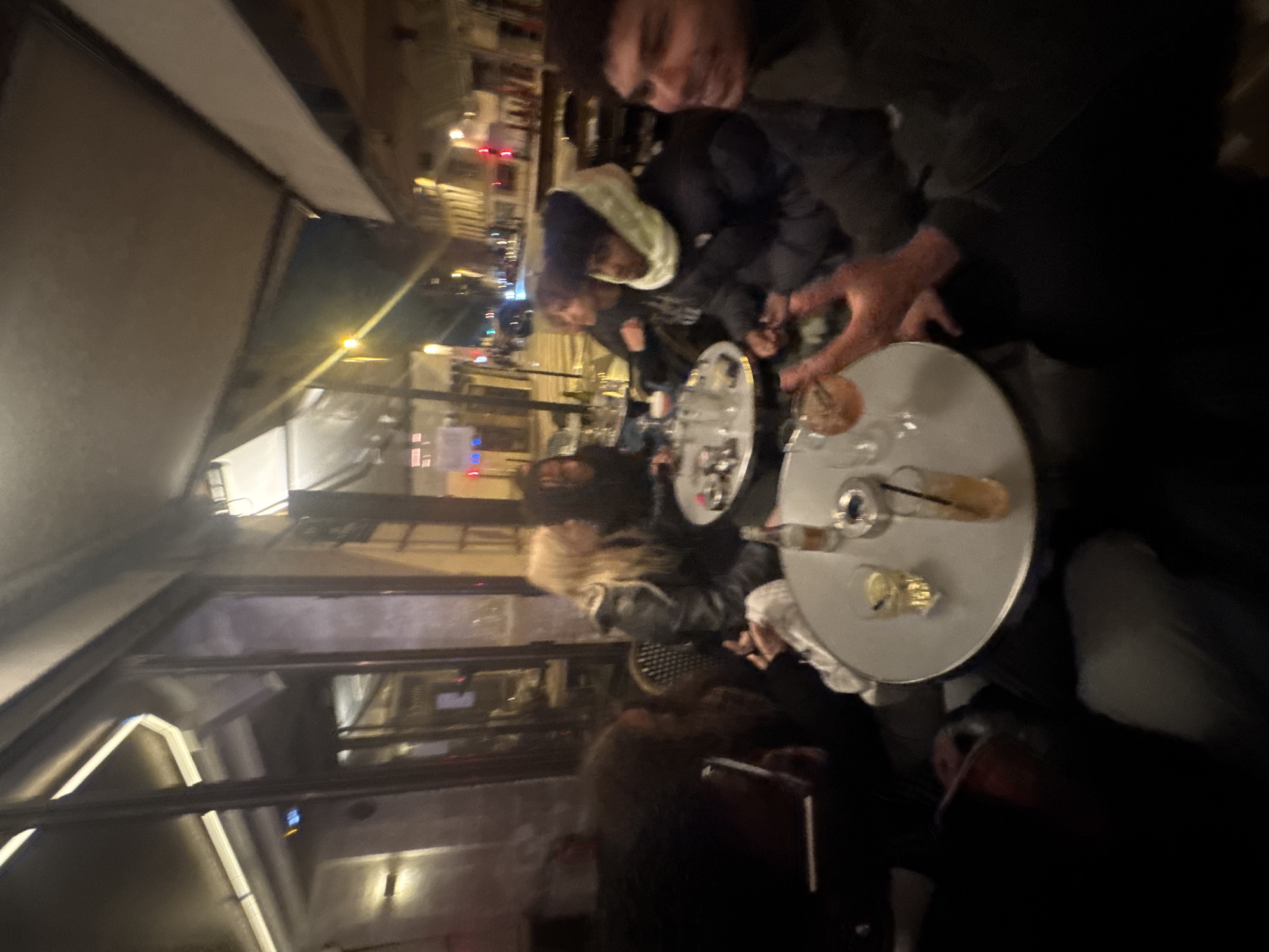 The image appears to be taken at an outdoor cafe in Paris, France, during the evening. Several people are seated around small, round tables, on which are drinks, possibly cocktails and beer, and ashtrays. The tables are placed close together. The foreground shows part of a window pane of the café, with reflections from the street outside. The background features a blurred cityscape with streetlights. The people are dressed in casual, dark clothing, suggesting it could be a cool evening. Several people are smoking.