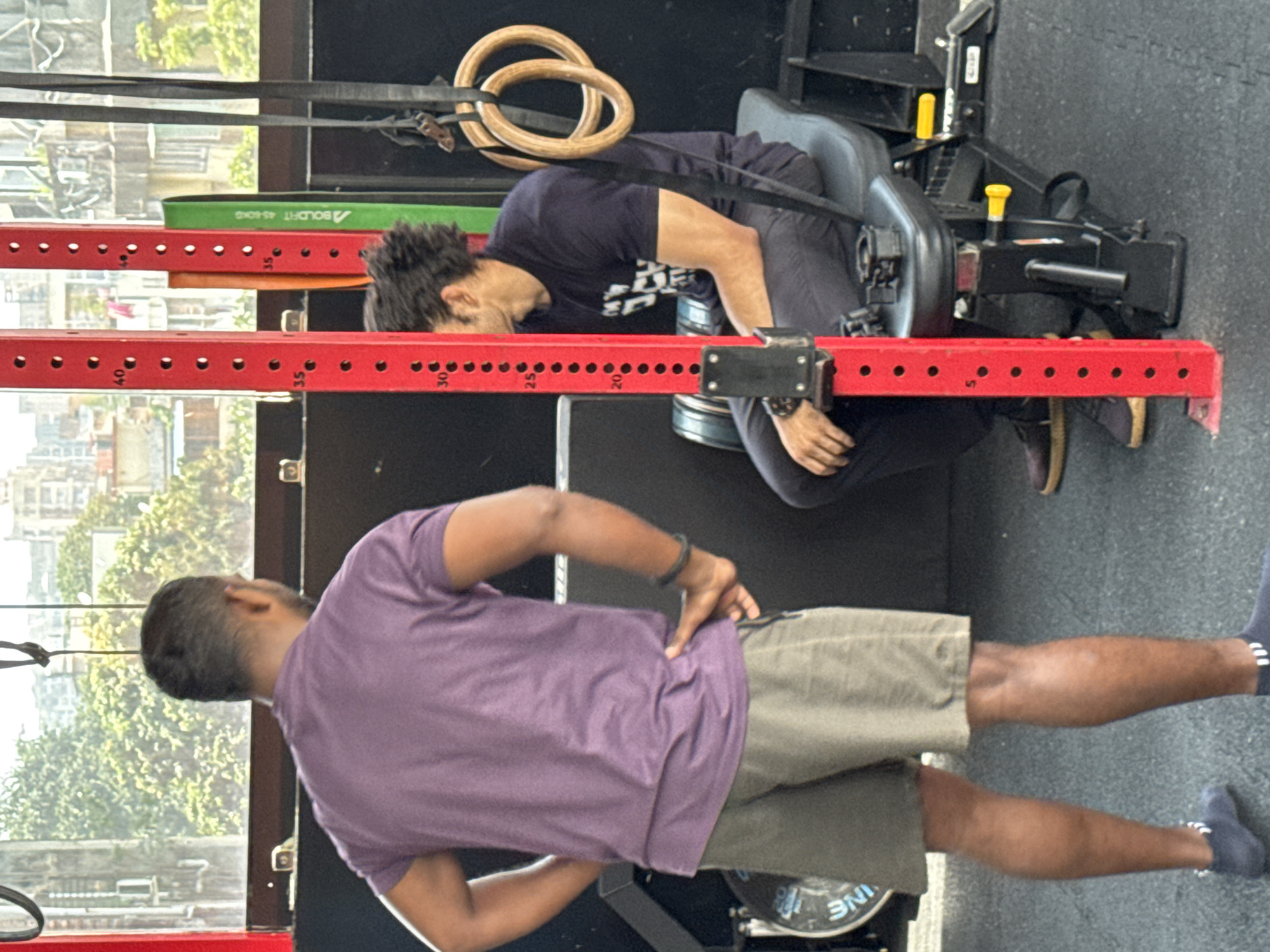 Two men are in a gym. One man is sitting in a leverage squat machine, resting his chest on the pad. He is wearing a dark blue t-shirt and dark pants. The other man is standing, wearing a purple t-shirt and khaki shorts, with his hand on his hip. The gym has red power rack uprights with numbered holes, and there are gymnastic rings hanging. The gym is likely located in Kolkata, India, judging by the buildings and foliage visible through the window.