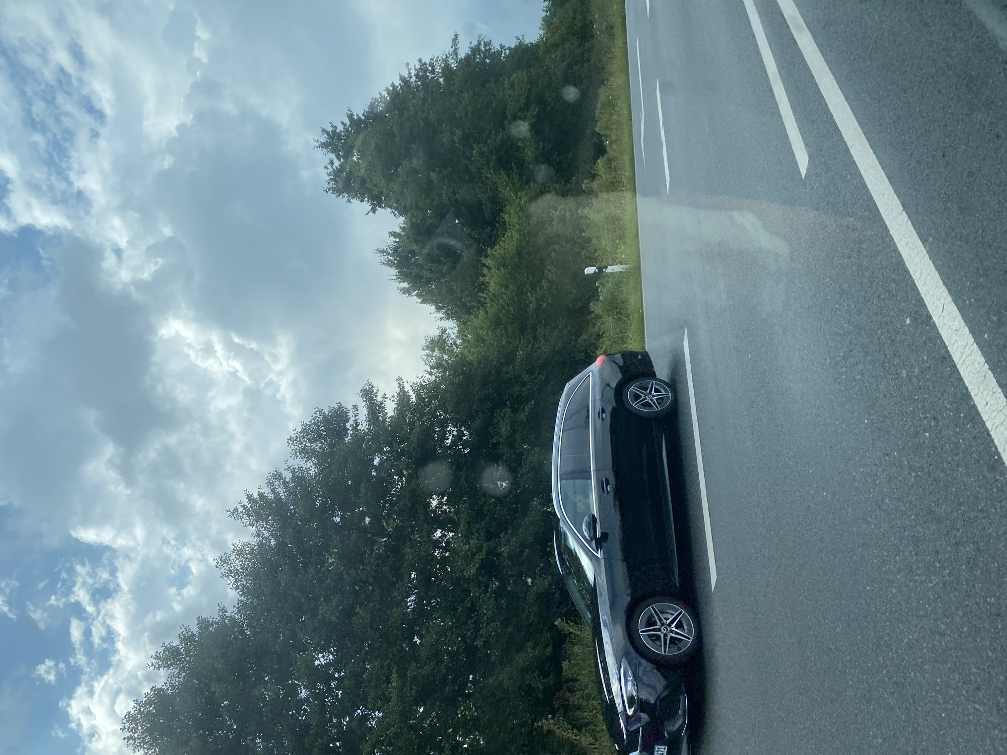 A black Mercedes-Benz C-Class Estate is driving on a wet road with white lane markings. The car is on the left side of the road, suggesting it is in a country where driving is on the right. The car has its headlights on, and the license plate is partially visible, appearing to be from Germany with "Bremen" and "HB" indicated. The environment is rural, with dense green trees lining the side of the road and a cloudy sky overhead. A white and red road marker is visible on the right side of the road. The image appears to have been taken from another vehicle moving in the same direction. The overall scene suggests a travel or commute on a damp day.