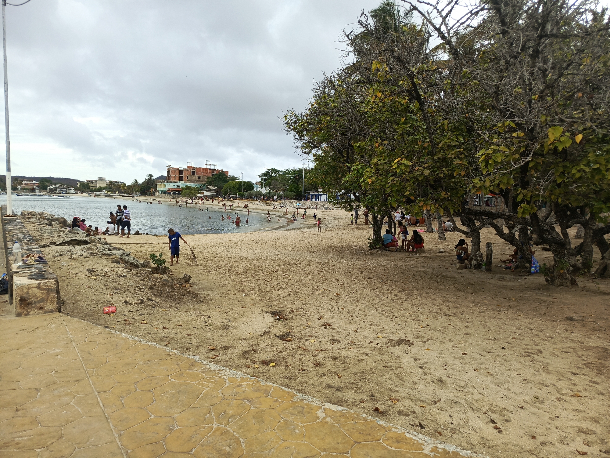 This image depicts a wide view of a beach or bay in Juan Griego, Venezuela, under an overcast, light grey sky. The foreground features a light brown, irregularly paved walkway and a rocky shore transitioning into a sandy beach. A white utility pole extends vertically on the far left.
In the mid-ground, numerous individuals are present. Many adults and children are wading or swimming in the calm, shallow water of the bay. On the immediate sandy shore to the left, a young person in a blue shirt holds a stick or branch, appearing to interact with the sand. Near them, two adult males in striped shirts stand at the water's edge. Further left, a small group of people are gathered on the rocky portion of the shore.
The background shows the far side of the bay with several small boats anchored or docked in the water. Beyond the water, various buildings are visible, including one prominent, multi-story brick structure that appears to be under construction or unfinished. Other residential or commercial buildings line the distant shore, interspersed with some vegetation.
The right side of the image is dominated by a cluster of trees, some with green and yellowing leaves, others with many bare, intertwining branches, providing shade over a substantial portion of the sandy beach. Beneath these trees, several groups of people are seated or lying on the sand, relaxing in the shade. The sand throughout the beach area is light brown and shows some darker patches, footprints, and scattered organic debris like leaves. A plastic water bottle and a red wrapper are visible on the sand near the paved walkway.