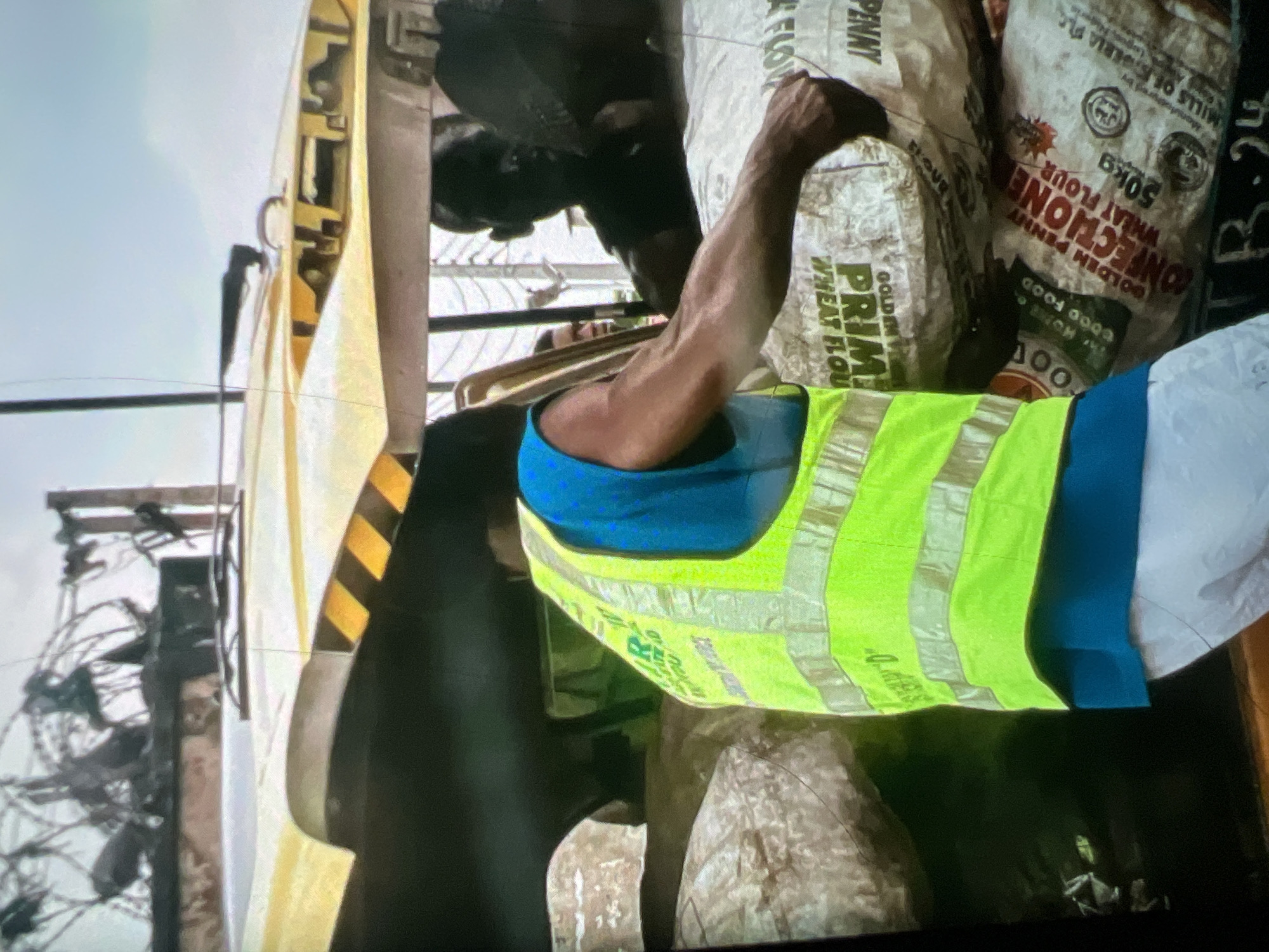 The image shows a man wearing a bright yellow-green safety vest over a blue shirt, standing next to a pile of large sacks. The sacks appear to be filled with flour, as evidenced by the text visible on them, which includes "PRIME WHEAT FLOUR" and "GOLDEN PENNY CONFECTIONERY WHEAT FLOUR 50KG". Some sacks also have Halal certification logos. The man's arm is extended, and he seems to be in the process of handling or arranging the sacks. In the background, a vehicle, possibly a three-wheeled auto-rickshaw or tuk-tuk, is visible, with a yellow and black striped canopy. The context suggests a street scene in Lagos, Nigeria, likely involving the transport or sale of goods. The environment appears to be outdoors, with a bright sky and some overhead wires and structures visible, typical of an urban setting in Lagos. Other people are partially visible in the reflection of the vehicle's surface, and within the vehicle itself. The overall impression is of a busy, everyday scene of commerce and labor in a Nigerian city.