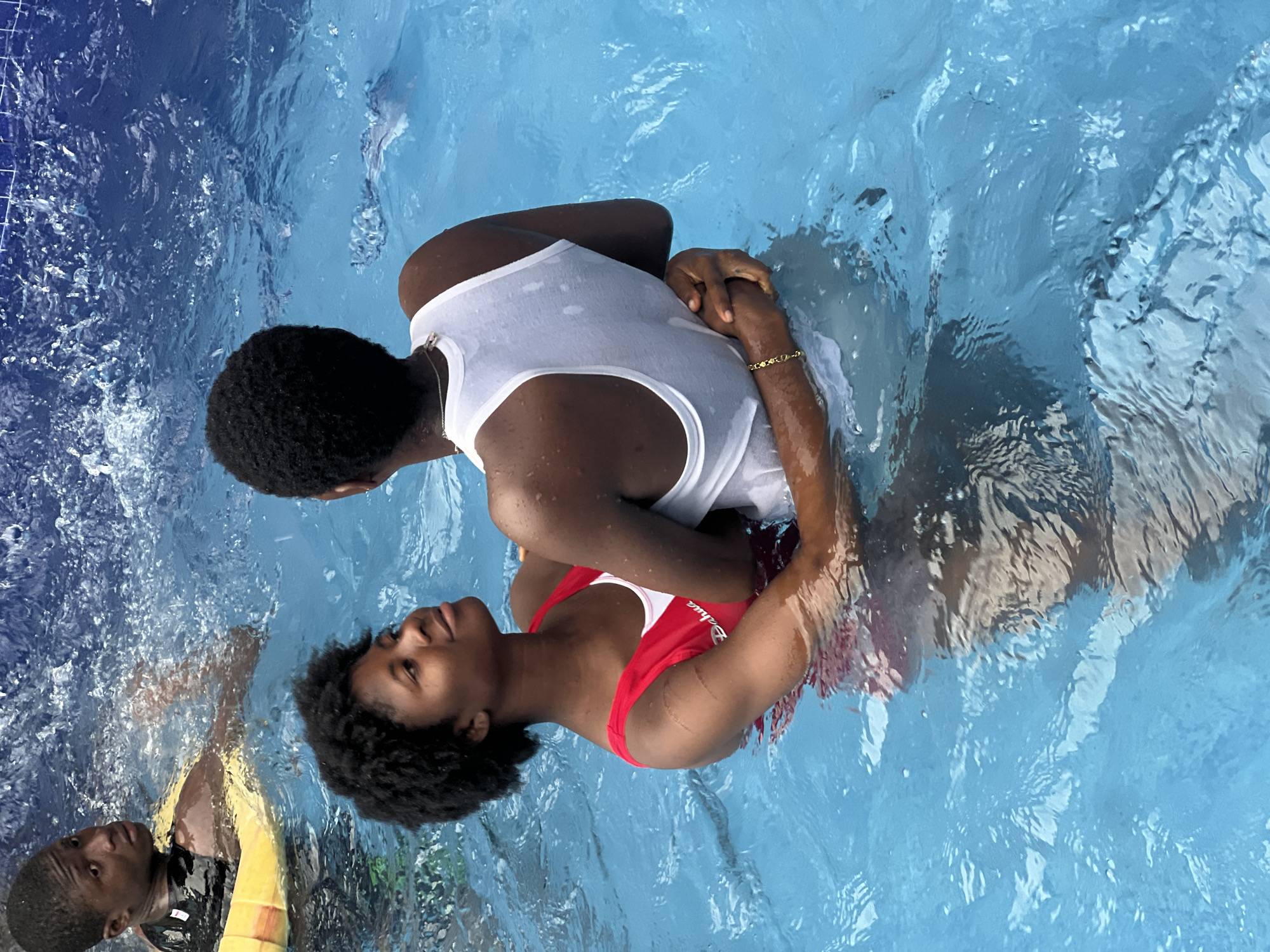 Two individuals are in a light blue swimming pool. One person is wearing a red swimsuit and is being held by another individual wearing a wet white tank top. The location is likely Obosi, Nigeria. Another individual is seen in the lower left corner of the frame wearing a yellow and black life vest.