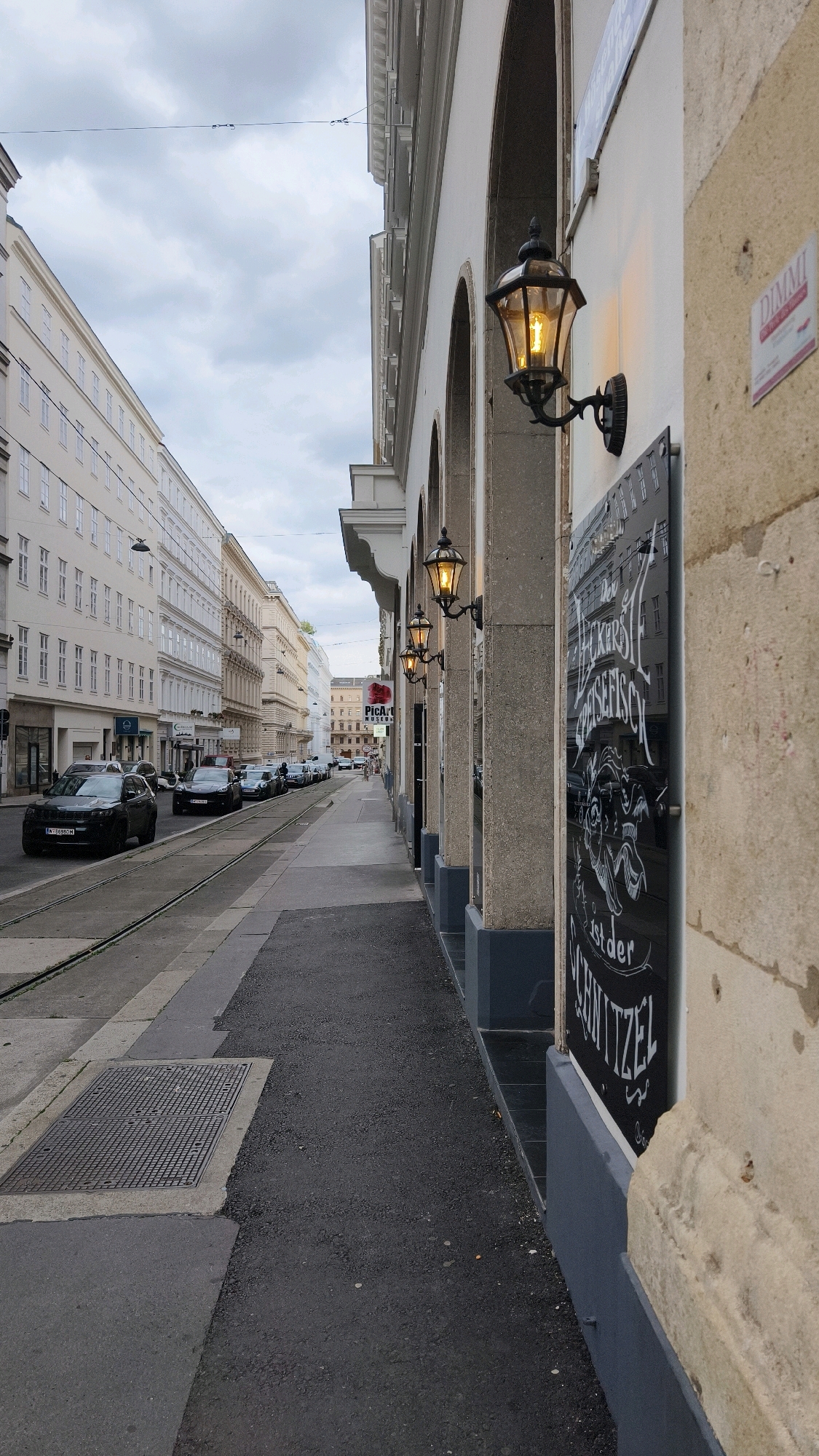 The image depicts a street scene in Vienna, Austria, likely a commercial area given the architecture and the presence of shops. The perspective is from the sidewalk, looking down a street lined with multi-story buildings. The sky is overcast, suggesting a cloudy day.
On the right side of the image, the facade of a building is prominent, featuring a series of arched openings. Mounted on this wall are two ornate, gas-powered street lamps, each emitting a warm yellow light. Below the lamps, a large black board with white lettering is affixed to the wall. The lettering appears to be a menu or advertisement, with prominent phrases like "Der Leckerste Speisefisch" (The Tastiest Fish Dish) and "ist der Schnitzel" (is the Schnitzel), indicating a restaurant. A small sign with the word "DIMMI" is also visible on the stone facade above the blackboard.
The sidewalk is paved with dark asphalt. To the left of the sidewalk are tram tracks running along the street. Several cars are parked along the street, and some are in motion. The buildings on the left side of the street are typical Viennese architecture, characterized by their light-colored facades and numerous windows. The street is relatively quiet, with no significant crowd of pedestrians visible. A manhole cover is set into the sidewalk near the tram tracks. In the distance, more buildings and a hint of a tram stop can be observed. The overall impression is of an urban environment in Vienna during daylight hours.