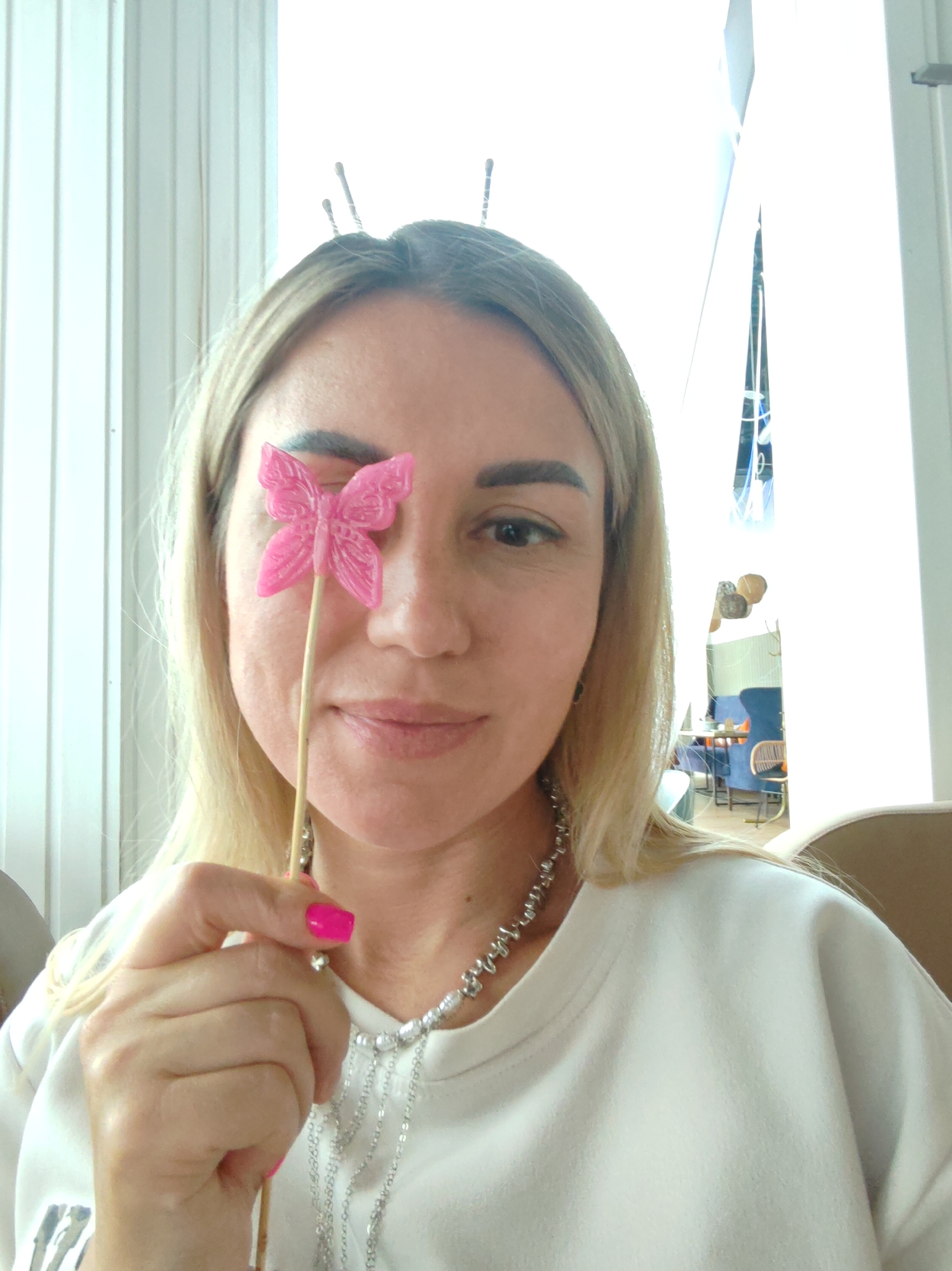A blonde woman is holding a pink butterfly on a stick in front of her left eye. She is wearing a white sweatshirt and a silver necklace. The background is blurred and appears to be an indoor setting with vertical blinds and some furniture. The image does not contain any information about the location being the city of Amursk, Russia.