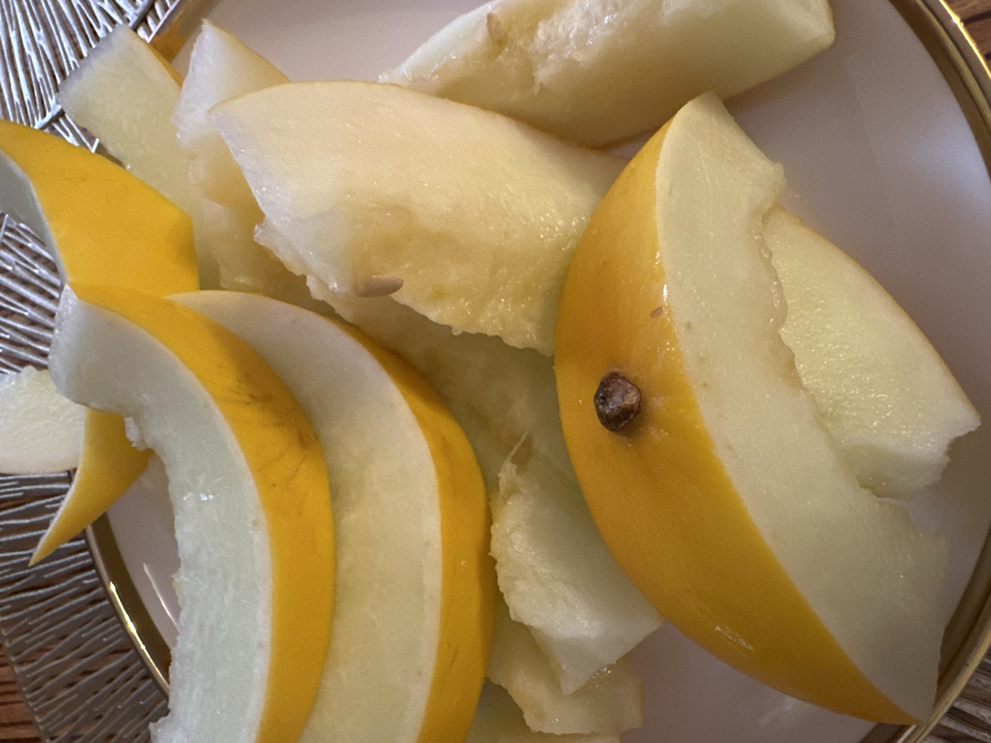 The image shows a close-up view of several slices of ripe honeydew melon served on a white plate with a gold rim. The melon is cut into wedges, with the rind being bright yellow and the flesh a pale creamy white. Some of the slices have visible seeds. The plate is set on a textured surface, possibly a placemat or tablecloth, with a reddish-brown and gold pattern. The lighting suggests an indoor setting, with a soft, diffused light source. There are no people or specific city landmarks visible in the image, and no information about Yuzhnouralsk, Russia is present.