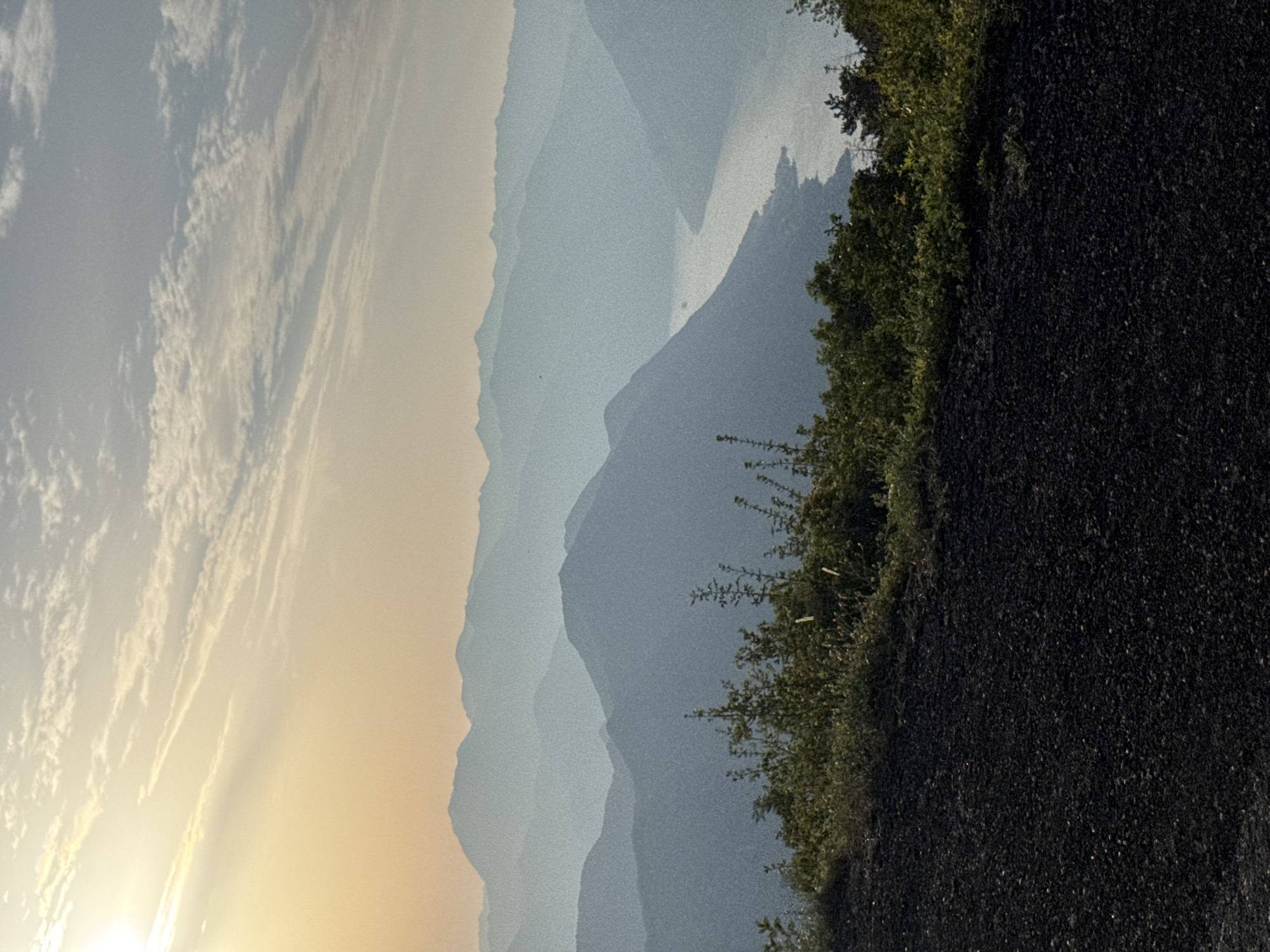 This image shows a panoramic view of a mountainous landscape, likely in the vicinity of Žanjev Do, Montenegro. The foreground consists of dark, rocky terrain covered with sparse, dry vegetation. Behind this, a series of lush green bushes and trees form a natural barrier.
Beyond the immediate foreground, the landscape unfolds into a dramatic vista of layered mountains. The mountains are shrouded in a soft, hazy blue light, with successive ranges appearing lighter and more distant, creating a sense of depth. A winding river or fjord is visible in the middle distance, reflecting the light of the sky. The water appears tranquil, with a small vessel navigating its course.
The sky is a mix of pale yellow and light blue, with scattered clouds. The sun, low on the horizon, casts a warm glow, illuminating the upper atmosphere and creating a serene, almost ethereal, ambiance. The overall impression is one of natural beauty and tranquility, with the vastness of the mountains and the gentle flow of water dominating the scene. The image is captured from an elevated position, offering a commanding view of the expansive terrain.