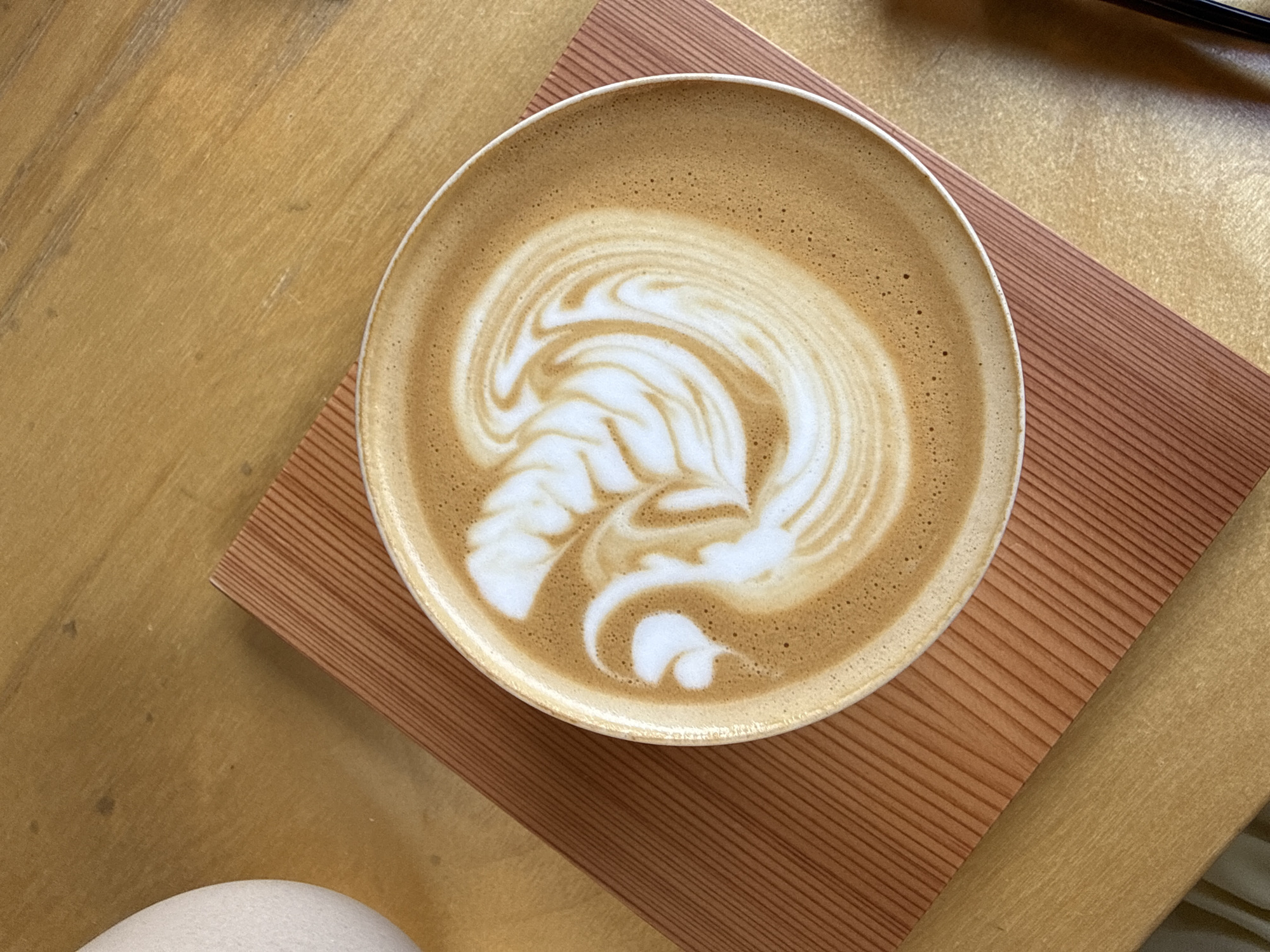 An overhead shot shows a latte with latte art in a light-colored ceramic bowl. The latte sits on a small, square wooden coaster with a grooved texture. The coaster and bowl are set on a light brown wooden table. A small portion of what seems to be a white mug or bowl is visible in the lower left corner. There is also a partial view of something dark (possibly a pen or utensil) on a different wooden surface in the upper right corner. No specific details suggest the scene is located in Tel-Aviv, Israel.