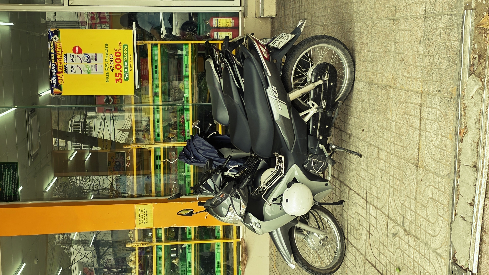 Several motorcycles are parked on a paved sidewalk in front of a storefront, likely a convenience store or small market, under bright daylight conditions. The motorcycles, primarily Honda Wave models and other scooters, are positioned closely together, facing towards the viewer's left. One light-grey scooter has a white helmet resting on its handlebars, and another has a blue bag or jacket draped over its seat. The ground is covered with patterned, light-colored square tiles.
The storefront features large glass windows, reflecting the outdoor environment including power lines, distant buildings, and possibly a person. Inside the store, green plastic crates filled with various items are stacked, and two red fire extinguishers are visible. Fluorescent lights illuminate the interior. A prominent yellow promotional poster is displayed on the inside of the glass, advertising a "GIÁ TIẾT KIỆM" (Saving Price) for P/S Procare toothpaste, offering a discount from 47,000 VND to 35,000 VND per tube, valid from June 1st to June 30th, 2026. The poster also mentions "SÔI ĐỘNG MÙA BÓNG" (Exciting Football Season).
A yellow vertical pillar marks part of the storefront entrance. On this pillar, a small yellow sign in Vietnamese reads: "QUÝ KHÁCH VUI LÒNG GIỮ TƯ TRANG CÁ NHÂN XIN CẢM ƠN" (Dear Customers, please kindly look after your personal belongings. Thank you). The overall scene, with the types of motorcycles, storefront, and Vietnamese text, is characteristic of a street in Vietnam, specifically Bac Nha Trang Ward.