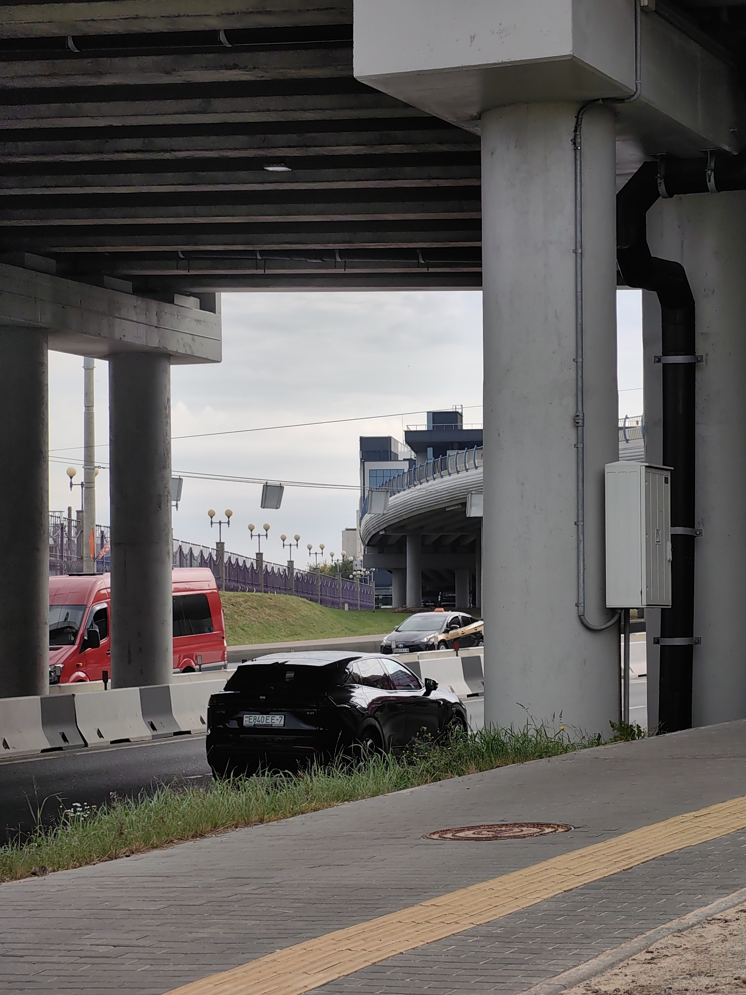 An outdoor, eye-level, medium shot captures a view of a black SUV parked on the side of a road, partially obscured by a concrete pillar. The SUV is facing right, with its rear visible. The license plate reads "E840EE-7". Behind the SUV, a yellow and black taxi is driving away on the road. A red van is parked on the left side of the road. In the background, a modern building with curved architecture is visible, as well as a pedestrian bridge with decorative railings and streetlights. The scene is set under an elevated highway structure, supported by thick concrete pillars. The ground in the foreground is paved with gray brick and features a textured yellow tactile paving strip. The sky is overcast. The location is in Minsk, Belarus.