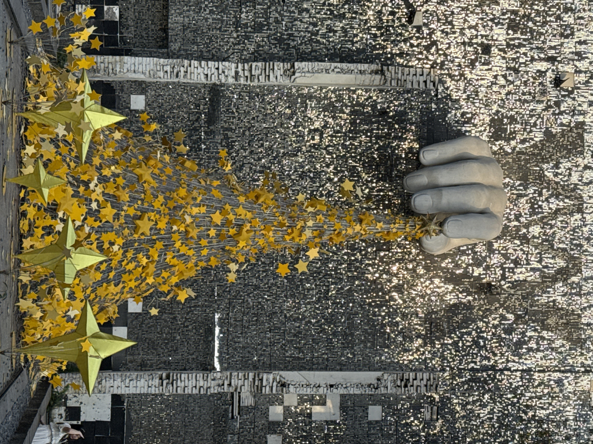 The image depicts a public art installation in Yuzhong District, Chongqing, China. A large concrete hand is positioned horizontally, appearing to release or grasp a stream of golden stars that emanate towards the left of the image. These stars are clustered and arranged to resemble a flowing, tree-like form, with larger, prominent golden star shapes positioned at the left-hand side. The background is a textured surface composed of repetitive dark and light geometric elements, suggestive of a building facade or public space. Some portions of the background glisten. At the bottom-left corner, two individuals are discernible. The scene is captured from a high angle, with the image rotated to present the main subjects horizontally.