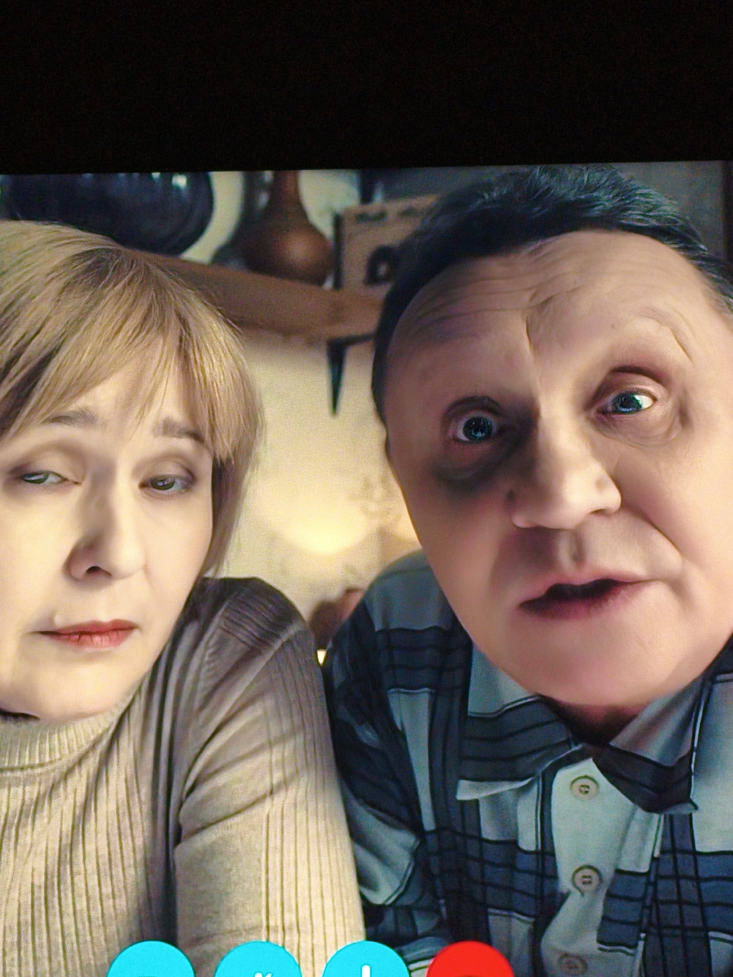 The image is a close-up, front-facing photograph of two people, a woman and a man, appearing on a screen, possibly a video call. The woman, on the left, has blonde hair and is wearing a light-colored ribbed sweater. She is looking slightly downwards and to her left. The man, on the right, has dark hair and is wearing a plaid shirt. His eyes are wide and directed towards the camera. The background is blurred, showing what appears to be a domestic setting with some decorative objects. There are also three circular icons at the bottom of the image: one blue, one white with a checkmark, and one red. There is no identifiable information in the image that would indicate a specific location in Vladivostok, Russia.