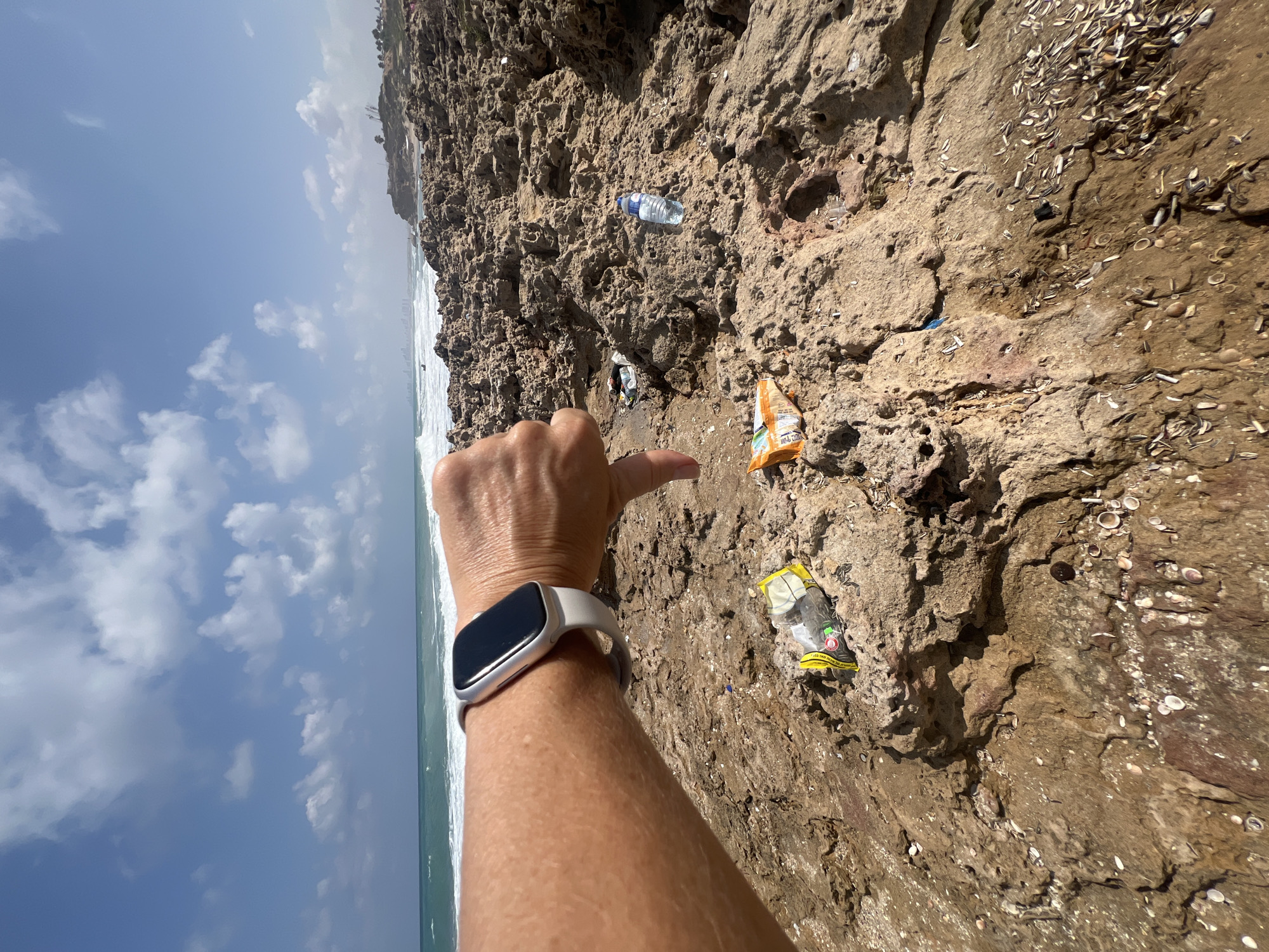 In Palmachim, Israel, a person wearing a white smartwatch on their left wrist gives a thumbs-down gesture. The scene is a rocky beach with a portion of the ocean visible in the background. Several pieces of trash are scattered on the rocks, including a plastic water bottle, a yellow and green chip bag, and other unidentifiable plastic debris. The sky is blue with scattered white clouds.