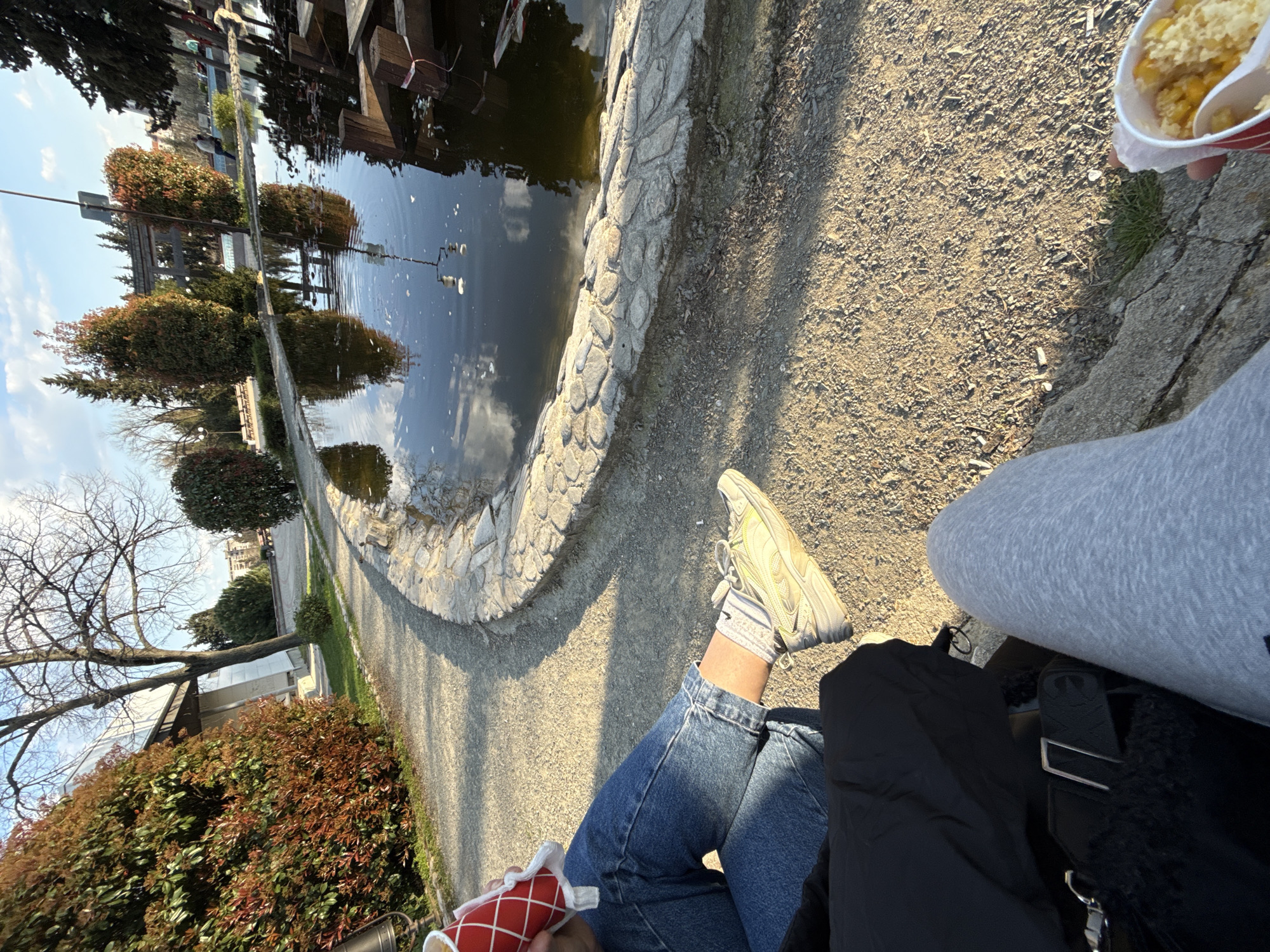 A person is sitting next to a pond at the park "Ayazmoto" in Stara Zagora, Bulgaria, with a container of food in hand. The pond has a stone edge. A walking path is visible next to the pond. The individual is wearing jeans and light-colored sneakers. A second person is sitting next to them wearing gray pants. A black jacket is visible, along with a bag.