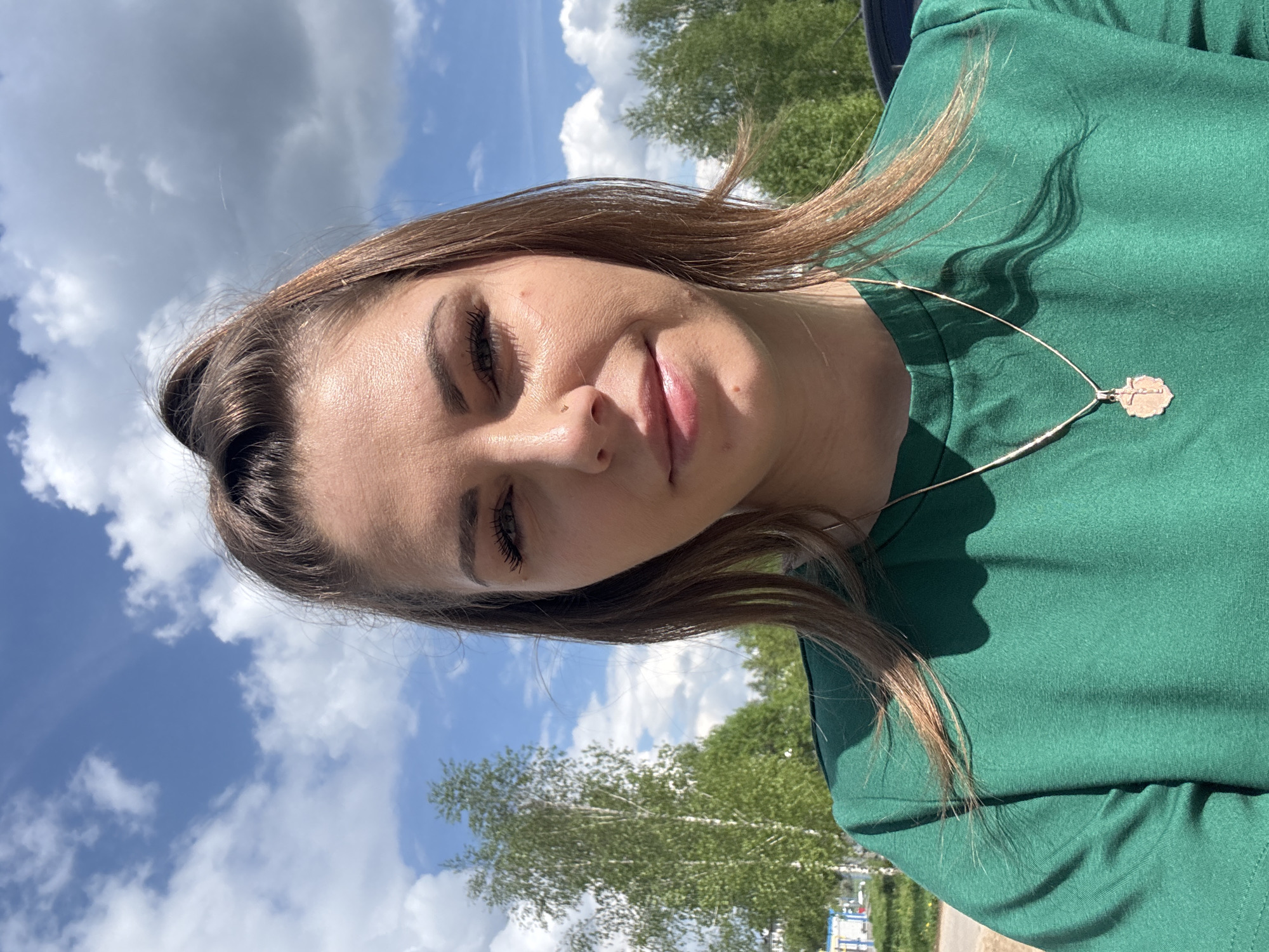 A young woman with brown hair is the primary subject of a selfie taken in an outdoor setting in Воронино, Russia. She is wearing a green shirt and a delicate necklace with a pendant. The background shows a partly cloudy sky and trees, suggesting the location is likely a park or green space within the city.