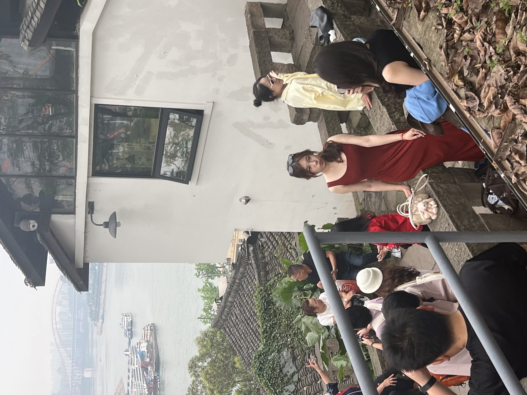 The image captures a scene from Nan'an District, China. The view includes the Yangtze River with several ships, a bridge, and buildings in the background. The foreground depicts a group of people on stone steps. Two women are posing for photos. One is wearing a yellow jacket and the other a red dress. There is a building with a window. There are dry leaves on the right side of the image. Other people are also on the steps, some looking at their phones, and others are standing by a grey metal railing. The weather appears overcast or hazy.