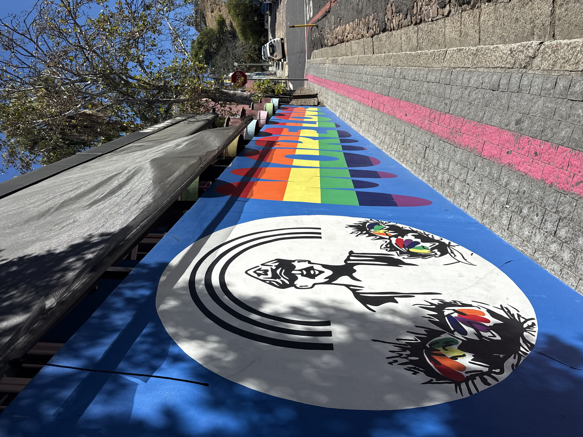 This image displays a brightly lit street scene in the Woodstock neighborhood of Cape Town, South Africa. A long, blue-painted wall runs along the left side, featuring two distinct murals. On the left, a large white circular area on the blue wall contains a black and white stylized illustration of a smiling individual wearing a peaked cap with a star emblem, positioned beneath a black triple-arc rainbow. Below this figure, two abstract, spiky shapes incorporate patches of rainbow colors. To the right, the wall is painted with horizontal rainbow stripes (red, orange, yellow, green, blue, purple) forming the words "LOVE WINS" in large, blocky letters. Above the rainbow mural, a series of inverted, pastel-colored buckets are attached to a ledge.
A grey cobblestone sidewalk with a prominent pink stripe painted down its center extends into the foreground. To the right of the sidewalk, a darker, rough-hewn stone border separates it from a paved street that slopes uphill into the distance. Several parked vehicles, including a white hatchback, are visible on the right side of the street. A red octagonal "STOP" sign on a pole is present in the middle ground on the right. A dark grey mesh awning extends over the wall, casting shadows onto its surface. Beyond the building and street, a vegetated hillside rises, with trees displaying green foliage against a clear blue sky. No people are visible in the scene.