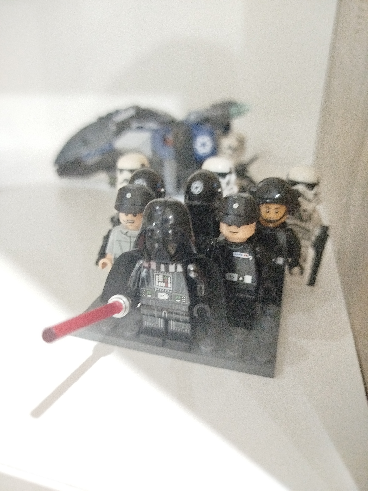 The image shows a collection of Star Wars Lego minifigures, including Darth Vader and several stormtroopers, arranged on a white surface. Darth Vader is in the foreground, holding a red lightsaber. Behind him are several other figures, some with white helmets and black armor, and others with black helmets. In the background, a blurry Lego spaceship is visible. The lighting is bright and diffused, suggesting an indoor setting. There is no discernible indication of the city of Mirny, Russia, or any other specific geographical location.