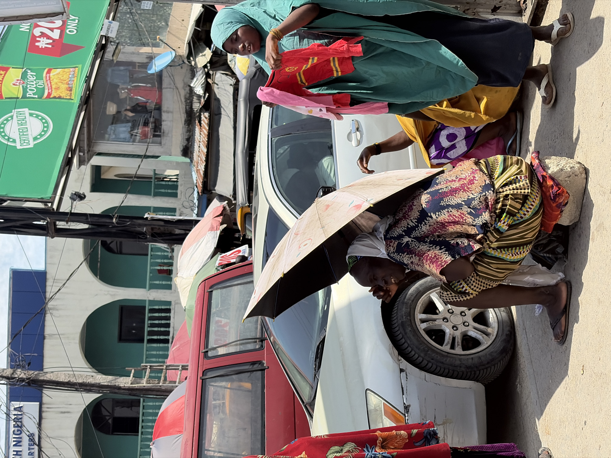 Here's a concise description of the scene:
The image captures a street scene in Lagos, Nigeria. Three women are prominent in the foreground. One stands holding clothes, while another is crouched under an umbrella next to a car tire. A third woman is partially visible standing next to them. They appear to be vendors or shoppers along the roadside. A red bus and several cars are also present. In the background, buildings with arched balconies and commercial signs are visible. One sign advertises "Power Oil".