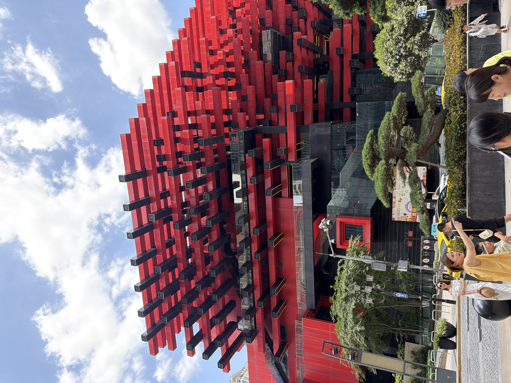 In Yuzhong District, China, a bright red and black modern building dominates the frame, its facade a complex arrangement of stacked red rectangular elements with black supports, jutting out at various angles against a blue sky with scattered white clouds. In the foreground, a paved walkway features several people. To the left, a woman in a white off-the-shoulder dress holds a straw hat. To her right, a woman in a yellow sleeveless top points towards the right of the frame. A young boy, partially visible, stands beside her. Further to the right, a man in a black t-shirt and shorts walks away from the camera. Behind them, lush green trees and bushes are visible, as well as streetlights, traffic signs, and yellow taxis on a street. The architectural style of the building is angular and geometric, with visible levels and windows. The overall scene depicts a bustling urban environment on a sunny day.
