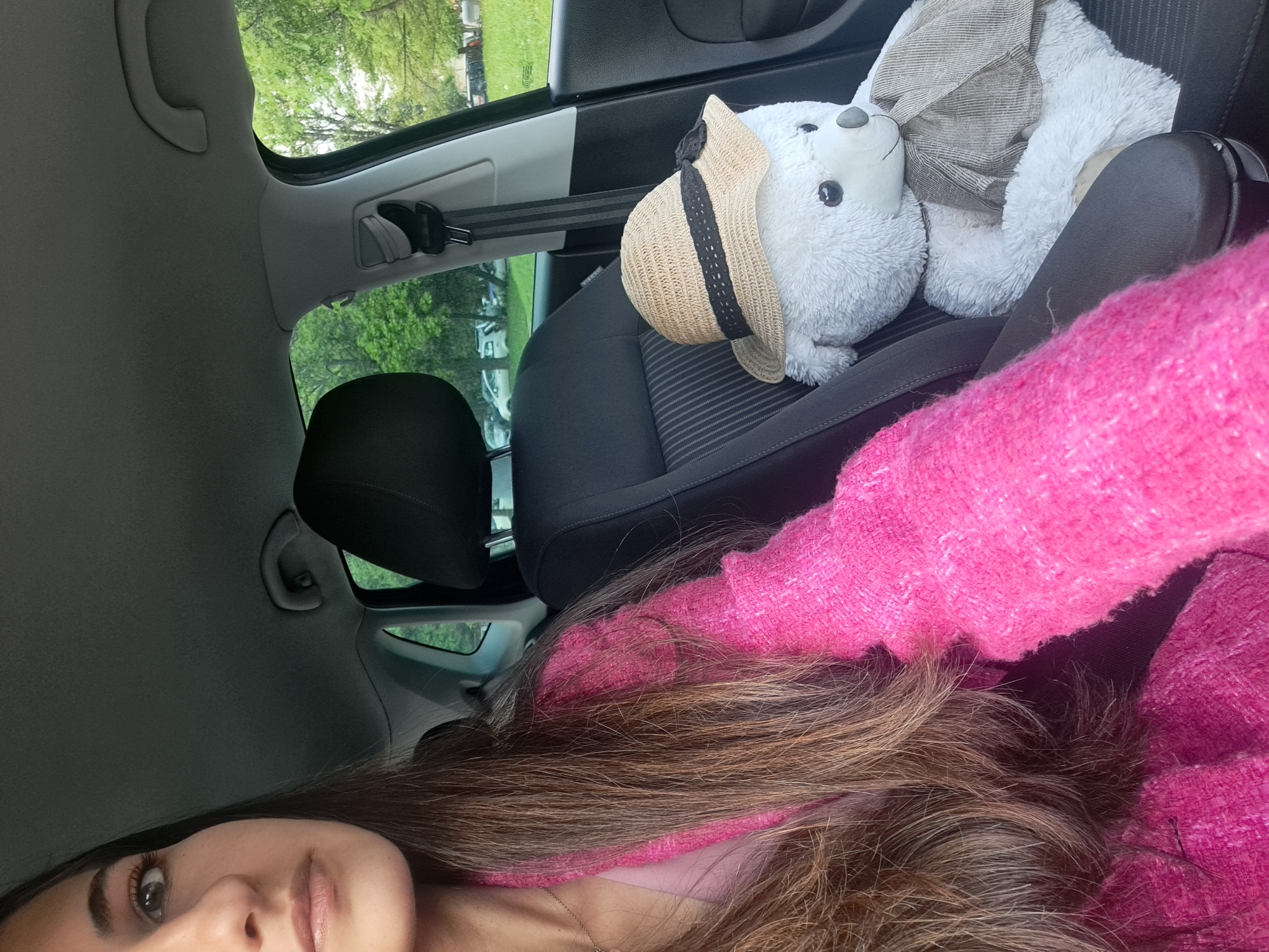 The image captures a selfie taken inside a car, likely in Sofia, Bulgaria. A person, visible from the nose upward, occupies the left side of the frame. She has long, dark brown hair and is wearing a vibrant pink, textured sweater. Her eyes are looking towards the camera.
To the right, on the passenger seat, sits a white plush toy resembling a seal or polar bear. The toy is accessorized with a light brown straw hat featuring a black band, and a grey striped scarf or blanket wrapped around its body. A seatbelt is fastened over the plush toy, securing it in place. The car's interior features dark grey fabric seats and a lighter grey headliner.
Through the car windows, a bright outdoor scene is visible, dominated by lush green trees. Partially visible parked white cars suggest an urban or suburban setting, possibly a parking area near nature. The natural lighting indicates it is daytime. No discernible text is visible within the image.