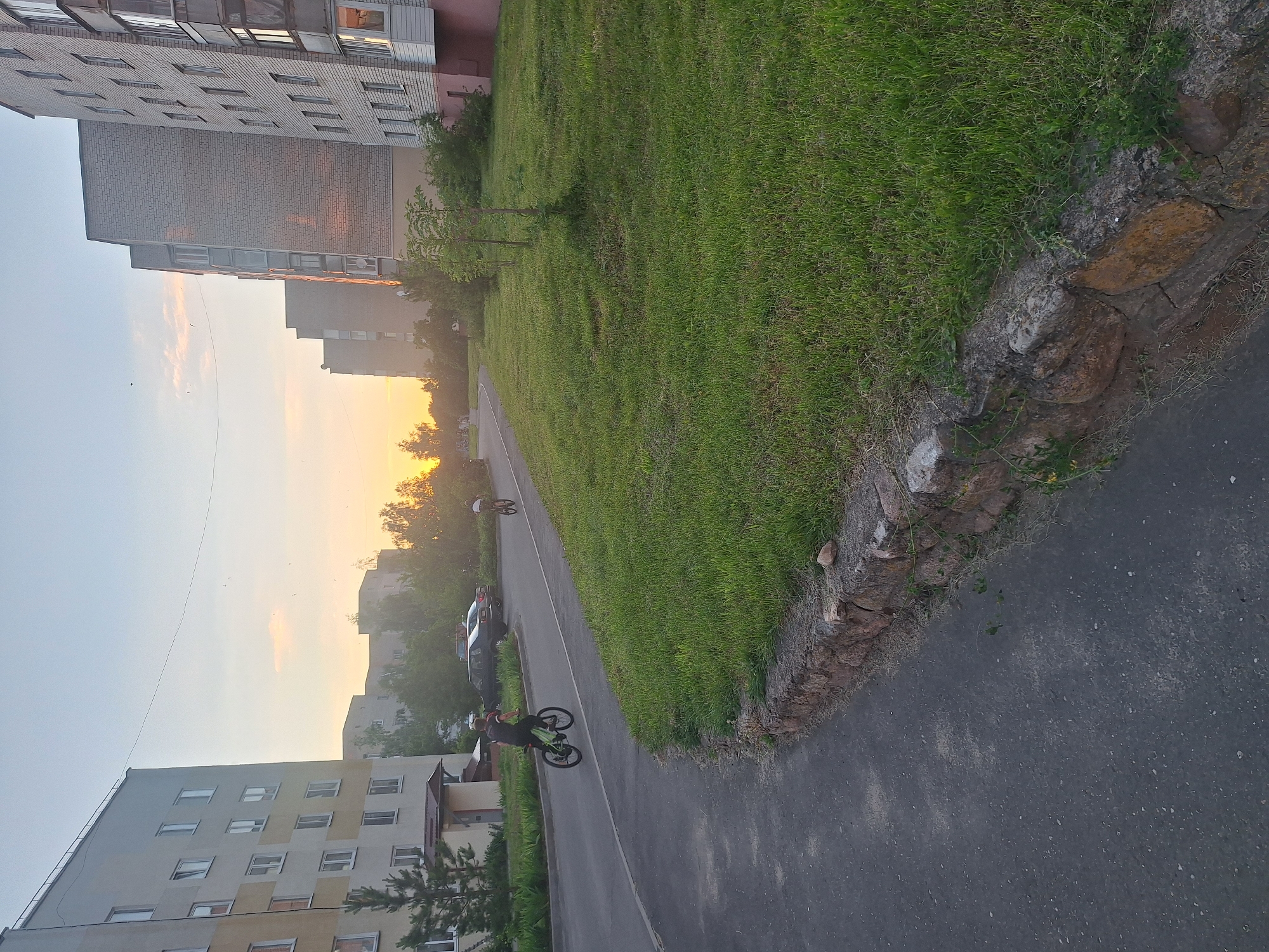 A wide shot captures a street in Капыльскі сельскі Савет, Belarus, during sunset. Two people are cycling on the asphalt road, with a young male on a green bicycle in the foreground and another individual further down the road. They are surrounded by residential buildings, including a multi-story apartment complex on the left and a brick building on the right. Lush green grass covers the area between the sidewalk and the buildings, bordered by a stone wall. The sky is hazy with the warm glow of the setting sun.