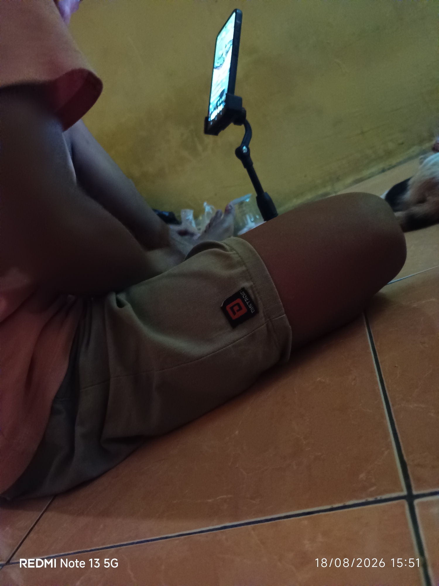A person is seated on the floor in what appears to be a domestic indoor environment. The person is wearing a light-colored t-shirt and khaki shorts. Their legs are crossed, and their hands are resting in their lap, partially obscured by their clothing. A smartphone, mounted on a flexible black stand, is positioned in front of the person and angled upwards, suggesting they are watching content on the device. The background consists of a yellow wall. A portion of a dog, primarily its head and back, is visible in the upper right corner of the frame, lying down. The floor is tiled with reddish-brown tiles. The scene is lit with artificial light, casting shadows. The timestamp "18/08/2026 15:51" and the text "REDMI Note 13 5G" are visible in the lower portion of the image, indicating the device used to capture the photograph and the date and time. The location in Talaga Sari, Indonesia, is not directly indicated by any visual cues in the image itself, but is provided as external context.
