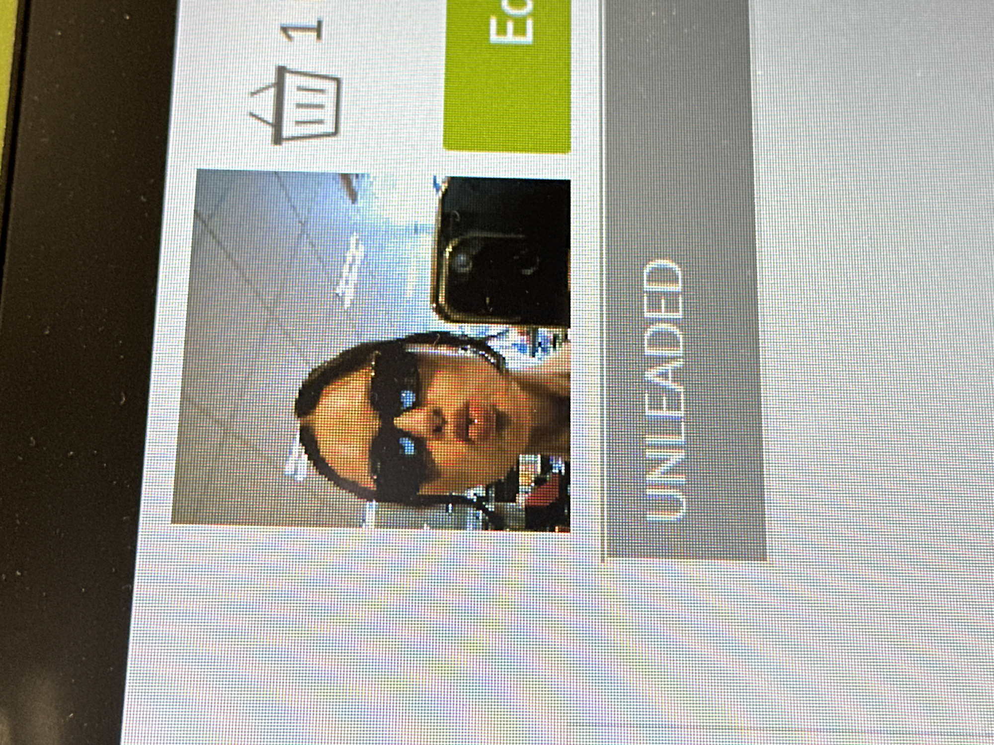 The image displays a close-up photograph of a digital screen, exhibiting a visible pixel grid. The screen primarily features a partially visible selfie-style portrait of a female. The female has dark hair, wears dark rectangular sunglasses, and her lips are pursed in a forward-facing 'kissing' gesture. A dark-colored smartphone is held to the right side of her face, partially obscuring her jawline and cheek, indicating it is the device used to capture the selfie. The background within the selfie suggests an indoor commercial or retail environment, with overhead fluorescent lighting fixtures and indistinct shelving units visible.
Additional digital user interface elements are present on the screen outside of the selfie. In the upper right corner, a shopping basket icon is displayed next to the numeral "1." To the right of the selfie, a partial green button with a white capital letter "E" is visible. Below the selfie, a grey horizontal bar contains the capitalized white text "UNLEADED." The image contains no definitive visual information to specifically identify its location as Tunbridge Wells, United Kingdom; the visible background elements are generic to retail settings.