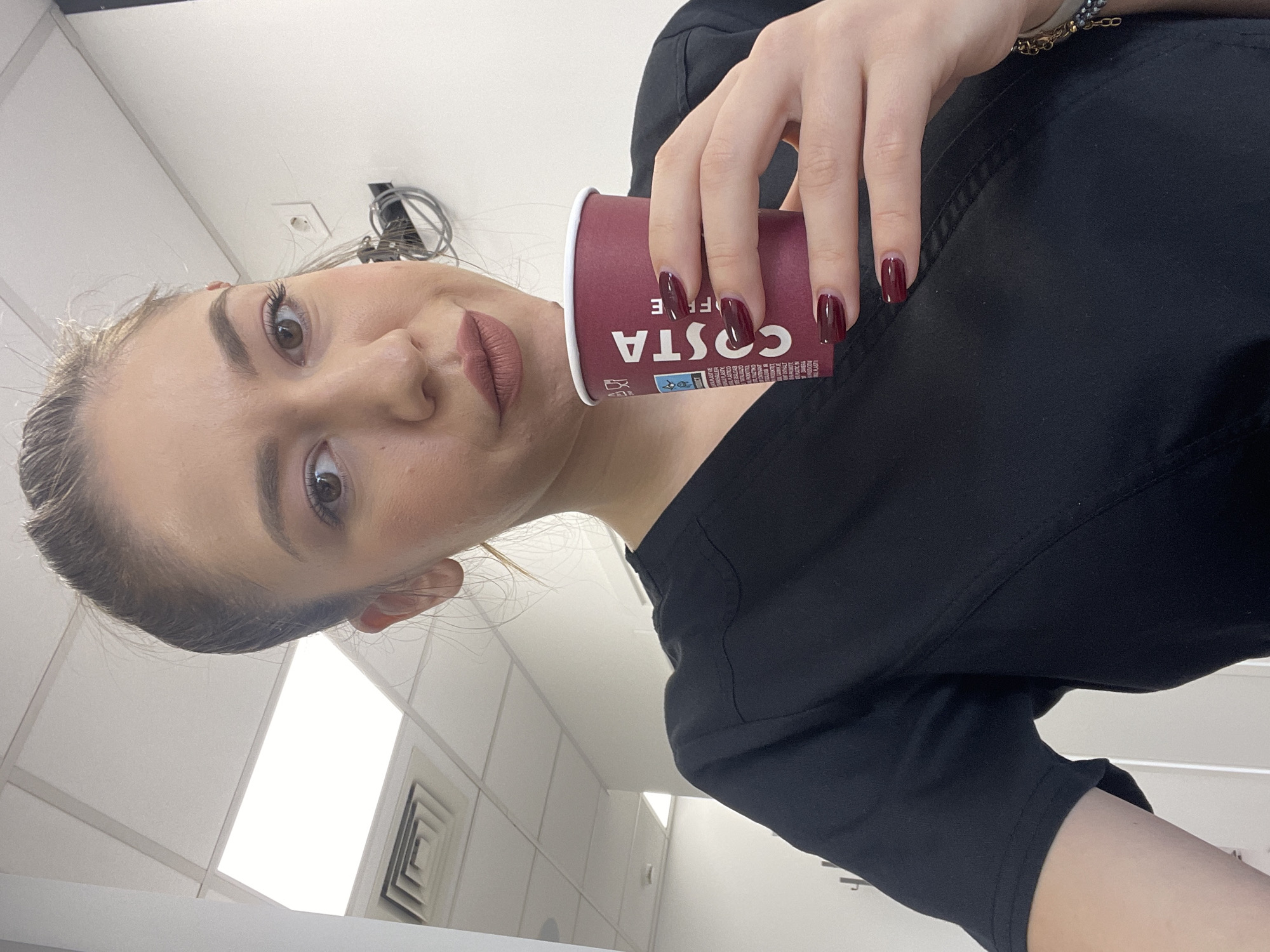 The image, taken in Sofia, Bulgaria, shows a young woman from a slightly low-angle perspective, holding a Costa Coffee cup. She is looking directly at the camera with a neutral to slightly smirking expression. Her brown hair is pulled back, and she has noticeable eye makeup, well-defined eyebrows, and dark, matte lipstick. Her fingernails are painted with dark red or maroon nail polish. She is wearing a black top or uniform, and a light-colored bracelet with gold-toned accents is visible on her right wrist.
The coffee cup, held in her right hand, is a branded Costa Coffee cup, predominantly maroon with the "COSTA COFFEE" logo visible, though inverted due to the image's orientation. Other smaller text and icons, likely related to branding or ingredients, are also present on the cup.
The background suggests an indoor setting, possibly an office, a commercial building, or a cafe. A white ceiling with square tiles, fluorescent lighting panels, and an air vent are visible. Some electrical wires are also seen attached to the ceiling. The lighting is artificial and bright. The image captures a casual moment, perhaps a coffee break.