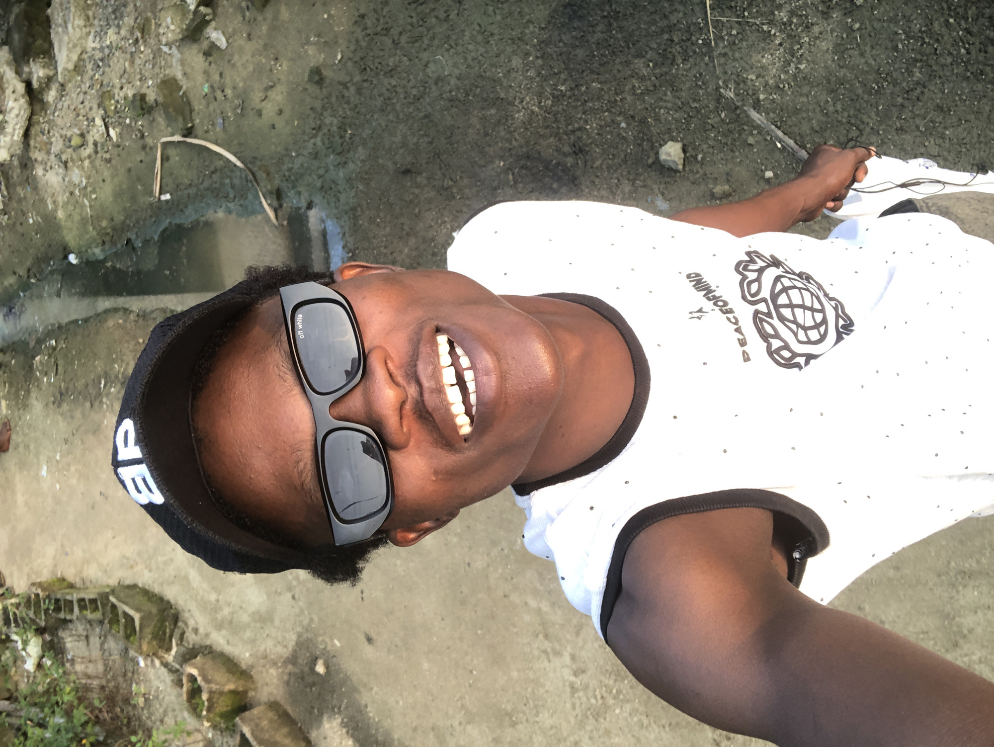 A man wearing a black baseball cap and sunglasses is taking a selfie outdoors. He has a bright smile, showing his teeth. He is wearing a white tank top with a design that includes the words "PEACEOFMINDF" and an illustration of a globe. The background appears to be an unpaved outdoor area, possibly a street or pathway, with some debris and a small ditch or channel of murky water. The overall setting suggests a simple, everyday environment.