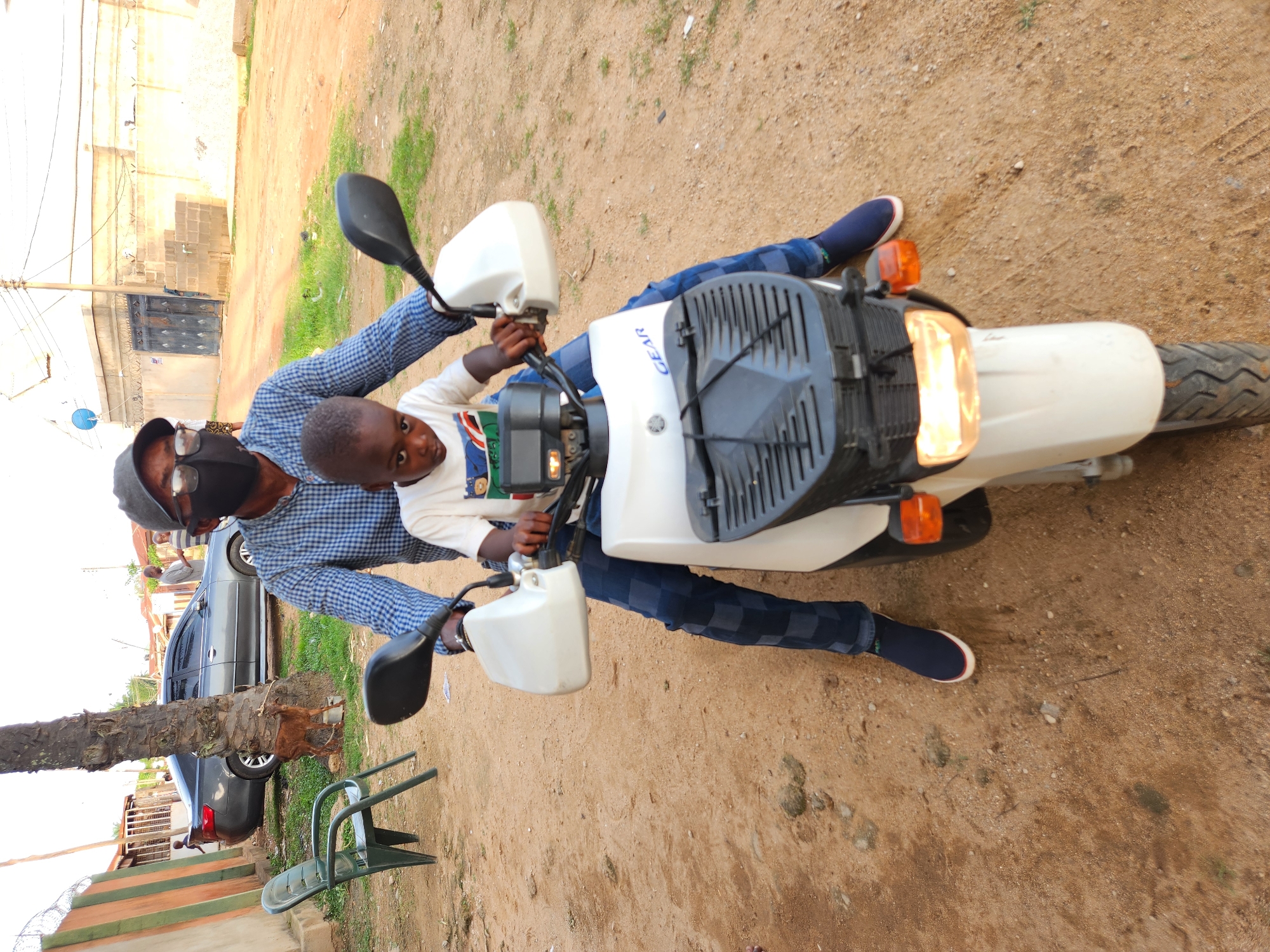 A man wearing a blue and white checkered long-sleeved shirt, dark pants with a similar checkered pattern, a grey baseball cap, black-rimmed glasses, and a black face mask is riding a white scooter. A young boy in a white t-shirt is seated in front of the man on the scooter. The man's left hand is on the handlebars, and his right hand appears to be resting on the boy. Both are facing forward, and the scooter's headlight is on. The scooter has a black basket in the front with a black wire cover. The scene takes place on a dirt road in Sabon Tasha, Nigeria. In the background, a grey sedan is parked to the left of a palm tree. A brown goat is tied to the palm tree with a rope. To the right of the road, there are buildings, including one with a light-colored facade and a dark metal gate, and a pile of cinder blocks. A green plastic chair is positioned to the left of the palm tree.