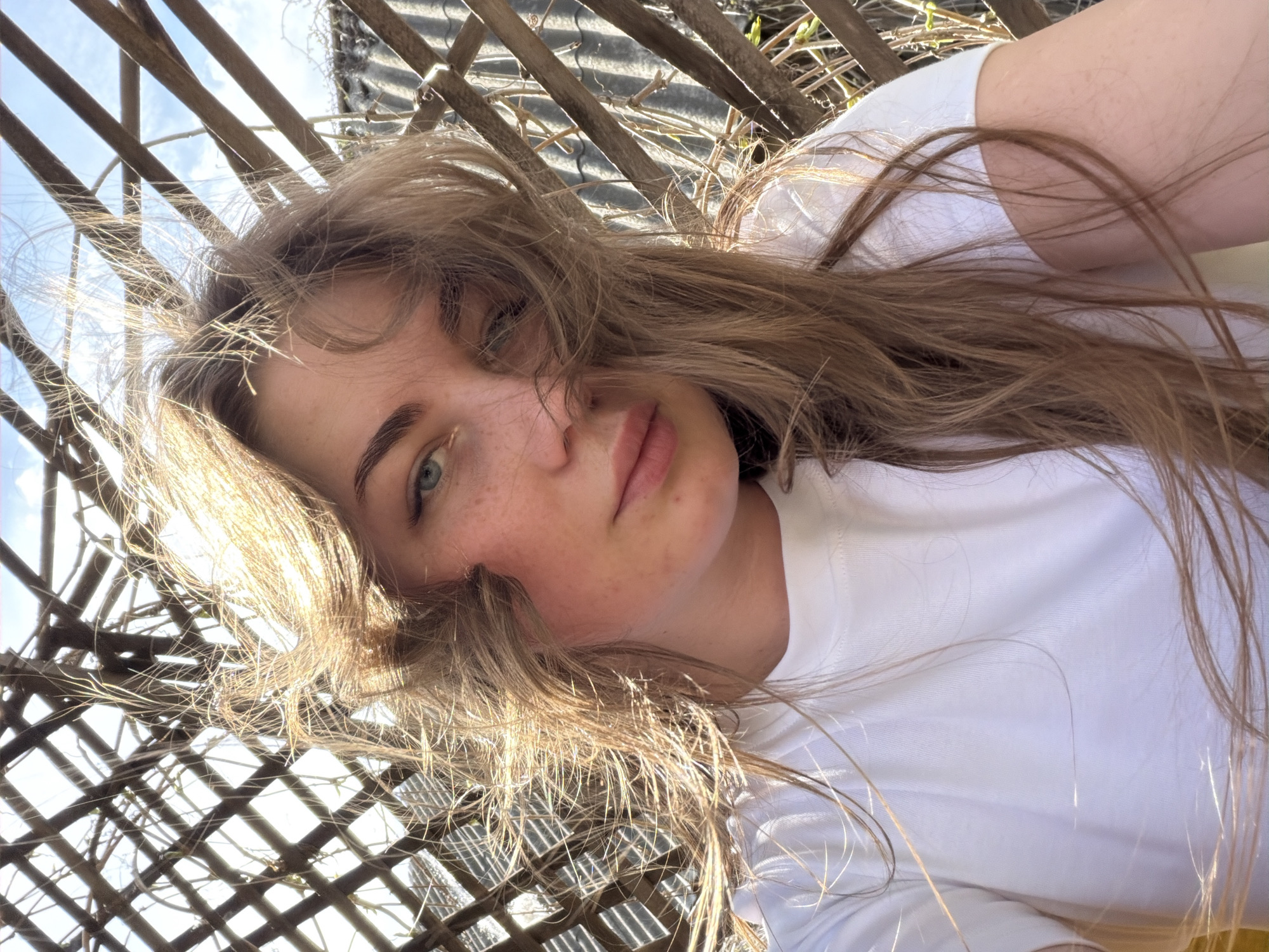 A young woman with long blonde hair and freckles is lying on her side, looking up at the camera. She is wearing a white t-shirt. Behind her, there is a lattice structure made of wood, with glimpses of the sky visible through the openings. The sunlight is bright and causes lens flare. The location is not discernible as being Уральское, Russia.