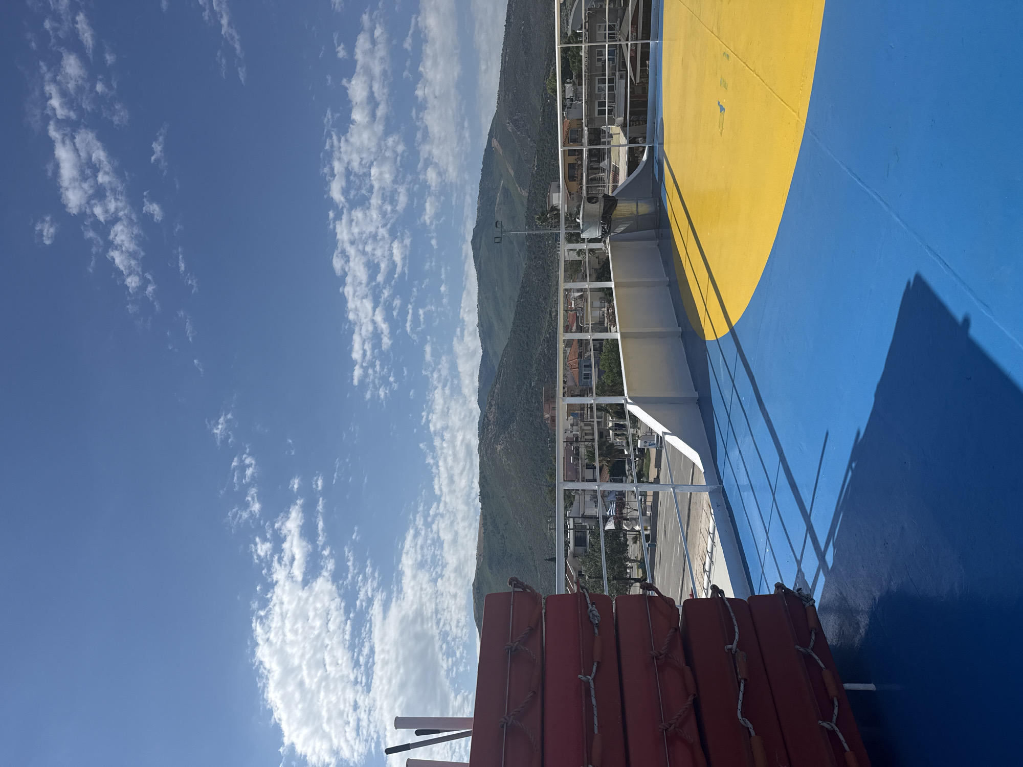 The image depicts a sunlit view from the deck of a ferry in Prinos, Greece. In the foreground, a stack of red life preservers is visible on the left, partially obscuring the blue and yellow painted deck. A white metal railing separates the deck from the background. Beyond the railing, a paved area with parked vehicles and buildings is visible. In the distance, a range of green, rolling hills forms the backdrop under a clear blue sky dotted with white clouds. The sun casts shadows on the deck, suggesting it is daytime.