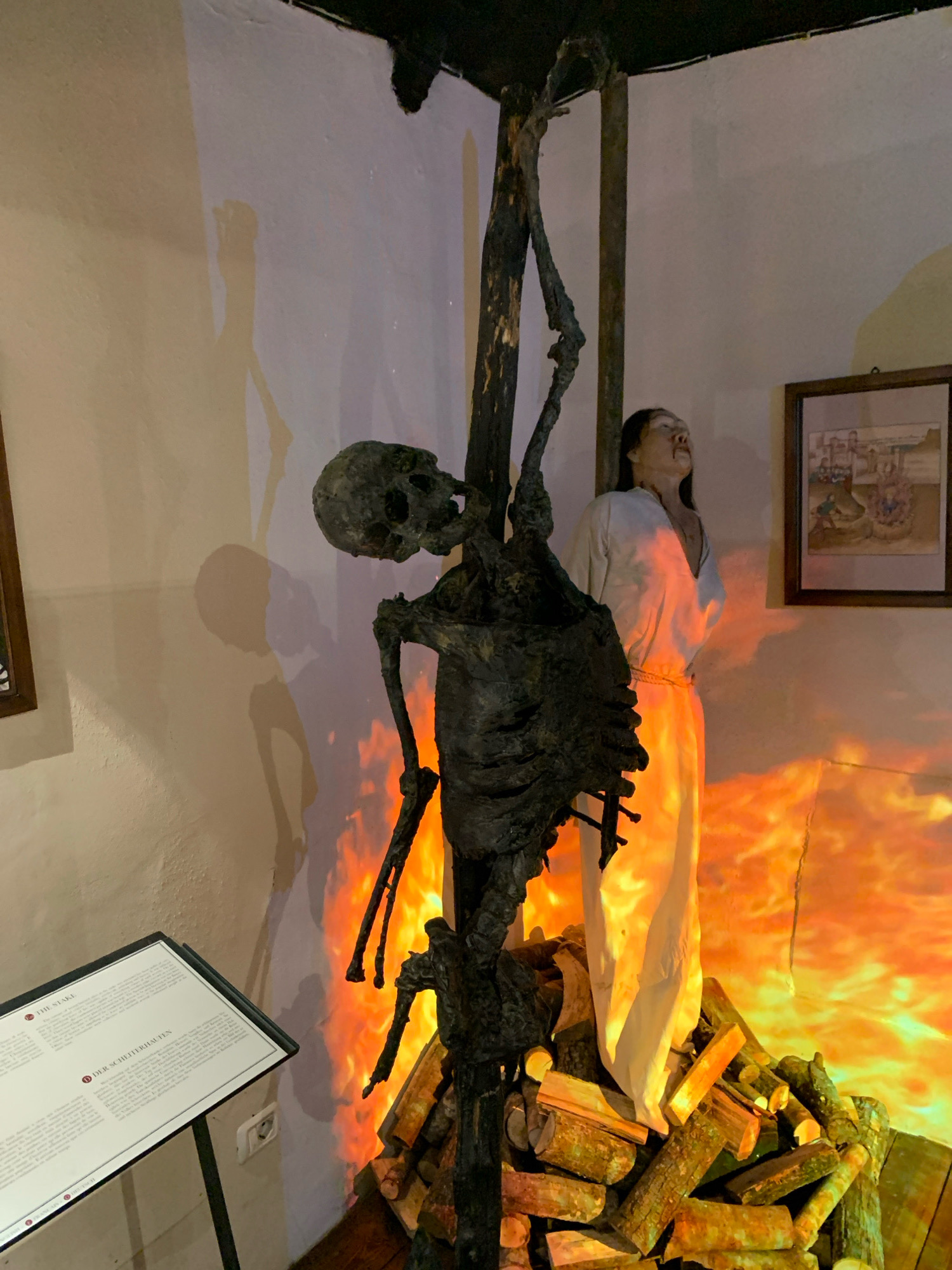 The image depicts a macabre diorama of a historical execution scene, likely from a museum exhibit, situated in the City of San Marino, San Marino. In the foreground, a decaying skeleton is suspended by ropes from a wooden stake. The skeleton is gaunt and appears to be made of dark, aged material. Flames, rendered in vibrant orange and yellow, are simulated at the base of the stake, surrounding a pile of chopped firewood, suggesting a burning at the stake. To the right of the skeleton, a life-sized mannequin of a woman stands draped in a simple white robe, her head tilted back and mouth open in a scream or gasp, as if enduring immense pain. She is positioned near the stake and the simulated fire. Behind the figures, the wall is painted a light beige. A framed picture hangs on the wall, showing a historical scene of multiple figures gathered around a similar execution. In the lower left corner, a display stand holds an informational plaque with text in English and German, providing context about the exhibit, with the visible titles "THE STAKE" and "DER SCHEITERHAUFEN." The lighting in the scene is dramatic, with spotlights highlighting the figures and the fiery backdrop, creating stark shadows on the wall. The floor appears to be wooden planks.