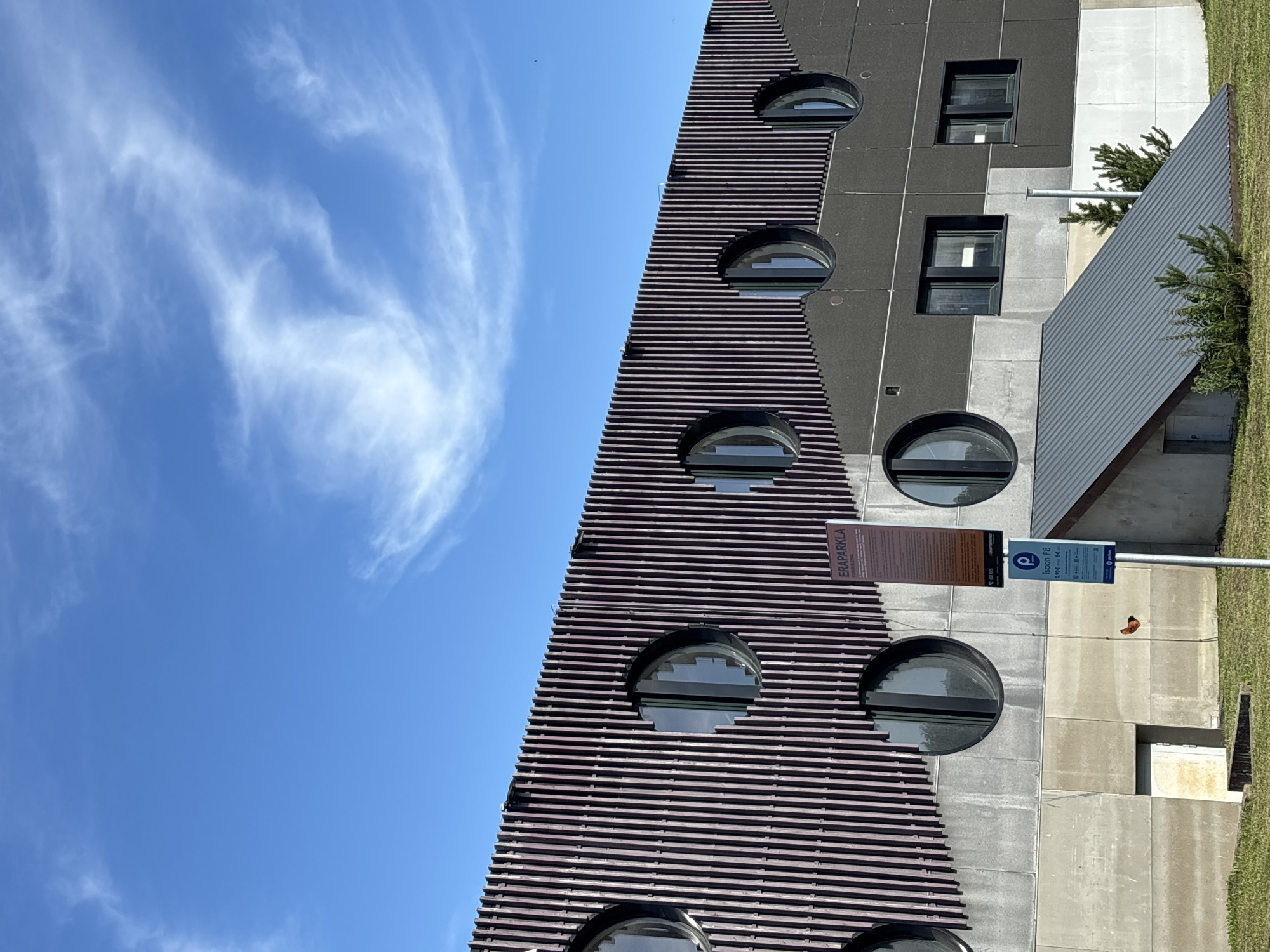 The image shows the modern facade of a building with a distinctive design in Haabneeme alevik, Estonia. The building features a dark, slatted upper section with purple undertones, punctuated by numerous circular windows. Below this, the facade transitions to a lighter gray concrete section, also with circular windows. Further down, there are rectangular windows and a sloping dark gray roof structure extending from the building, partially covered in grass and small evergreen trees. Two information signs on poles are visible in front of the building. The sky is a clear blue with wispy white clouds. The overall scene conveys a contemporary architectural style against a bright, daytime sky.