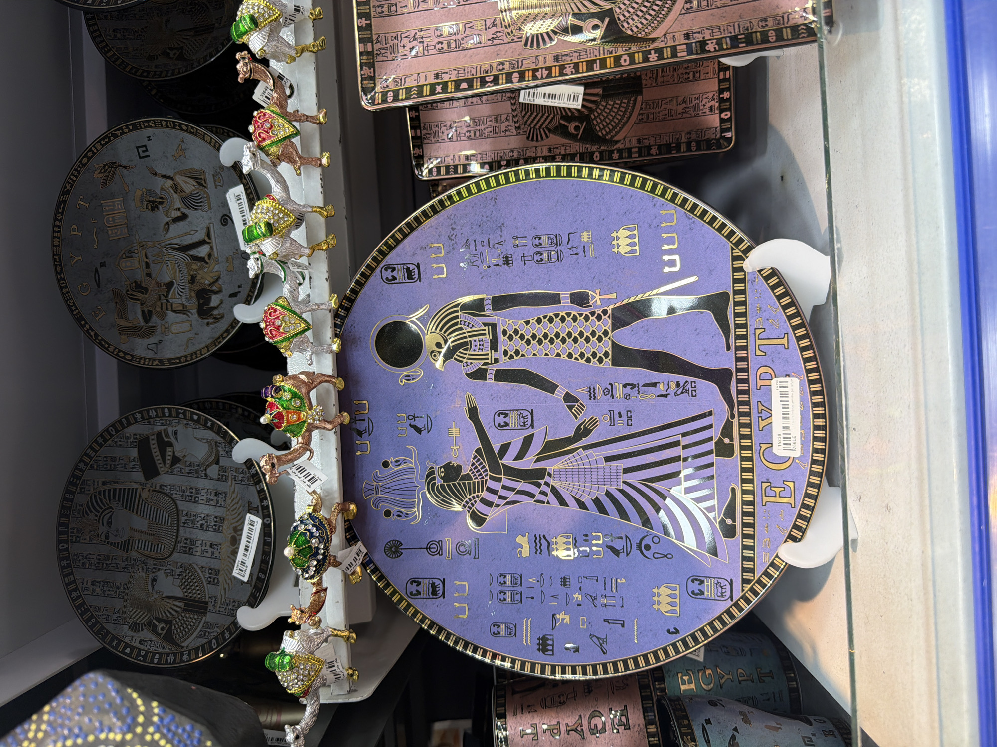 In Sharm El Sheikh, Egypt, a display shelf showcases Egyptian-themed souvenirs. Prominently featured is a large, round, purple plate adorned with golden hieroglyphs and depictions of Egyptian deities and figures. Several smaller, jewel-encrusted camel figurines stand in front of the plate, each decorated with vibrant colors and metallic accents. Behind the large plate, other Egyptian-themed decorative plates are visible, some featuring detailed hieroglyphic patterns and portraits of pharaohs. To the right and left of the main display are rectangular items, possibly boxes or trays, also decorated with Egyptian motifs and hieroglyphs. The overall scene suggests a retail environment where these items are for sale, likely in a shop catering to tourists in Sharm El Sheikh.