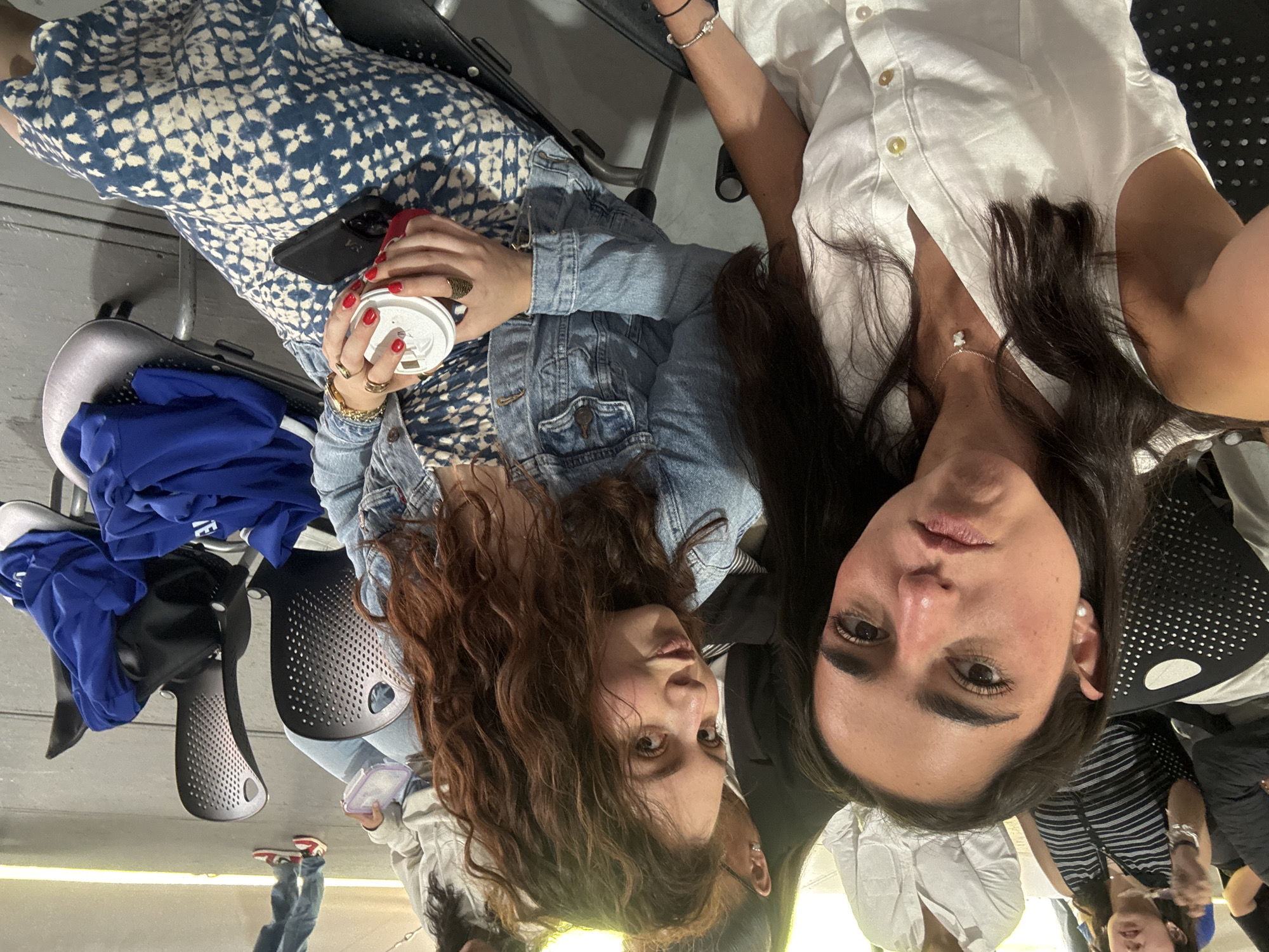 Two women are featured prominently in the foreground of this indoor scene, looking directly towards the camera. The woman on the left has long, wavy reddish-brown hair and is wearing a denim jacket over a blue and white patterned dress or skirt. She holds a red cup with a white lid in her hands, which are adorned with several rings. A black smartphone, featuring a "VS" logo on its case, rests on her lap. To her left, a blue garment is casually placed on an empty black chair.
The woman on the right has long, dark brown hair and is dressed in a white button-down shirt. She wears a delicate silver bracelet on her left wrist and a necklace with a small pendant. Pearl-like stud earrings are visible on her ears.
They appear to be seated in an area with multiple black chairs, some with perforated backs and seats, suggesting a waiting room, event space, or similar public indoor venue. The floor is light-colored. In the background, other individuals are partially visible, with one person wearing red and white sneakers. The overall lighting suggests an indoor setting. Both women have neutral expressions, posing for the photograph.