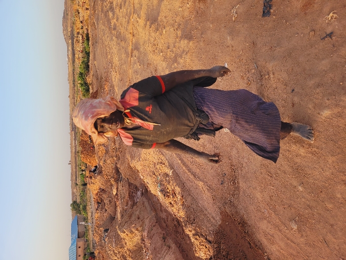 The scene is an outdoor, arid landscape in the city of Shen, Nigeria, characterized by reddish-brown earth, indicative of a disturbed area such as a quarry, brick-making site, or construction zone.
**Central Figure:** A dark-skinned individual, likely female, walks on an unpaved dirt path, moving towards the viewer. The individual is barefoot, and their skin, particularly on the arms and lower legs, is coated in reddish-brown dust. They wear a dark grey short-sleeved collared shirt with red shoulder and placket accents. A light-colored fabric, possibly faded pink, covers their head, draped to form a wide brim around the face. A dark skirt with faint vertical stripes extends below the knees.
**Environment and Background:** The ground is uneven, comprised of dry, compacted reddish-brown soil, with numerous excavated piles and depressions. In the mid-ground to the left, a single-story structure with reddish-brown walls and a light blue roof is visible, adjacent to a smaller, light-colored building. Distant, indistinct figures are scattered among earth mounds in the far background. Sparse green vegetation is visible beyond the immediate disturbed area. The sky is clear and light blue, suggesting fair weather. The overall lighting indicates either early morning or late afternoon.