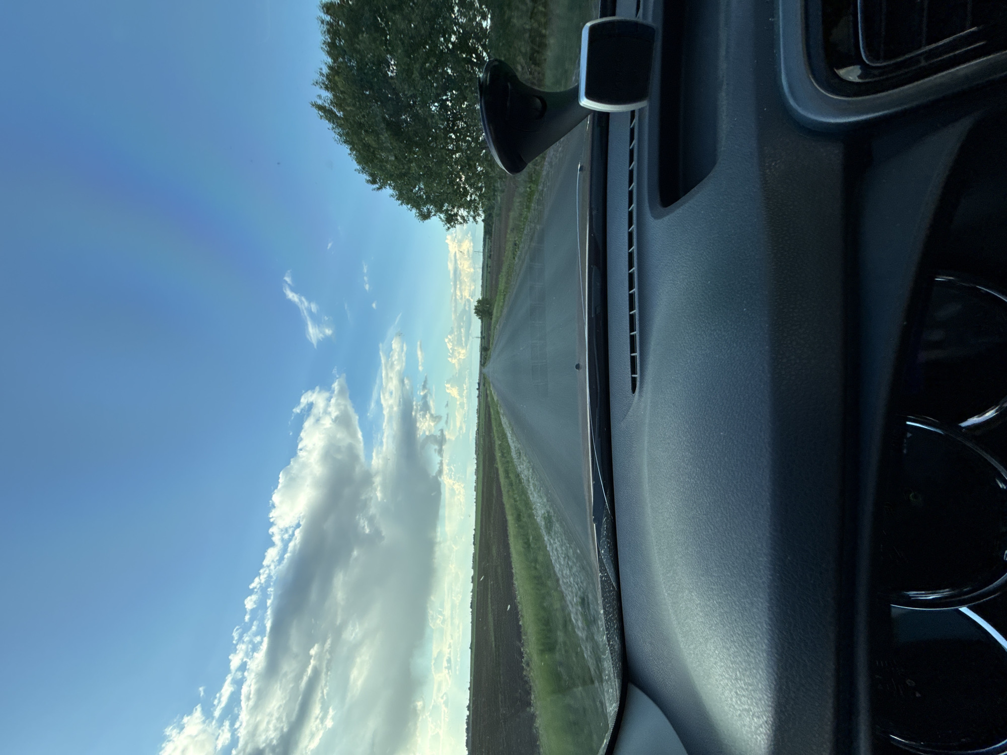 The image shows a view from inside a vehicle driving on a paved road. The road stretches into the distance towards a horizon, flanked by fields on either side. A tree is visible to the left of the road. The sky is blue with scattered clouds. A car dashboard is visible on the right side of the image. The location cannot be determined to be Merinovshchina, Russia based on the information provided in the image. There are no people visible.