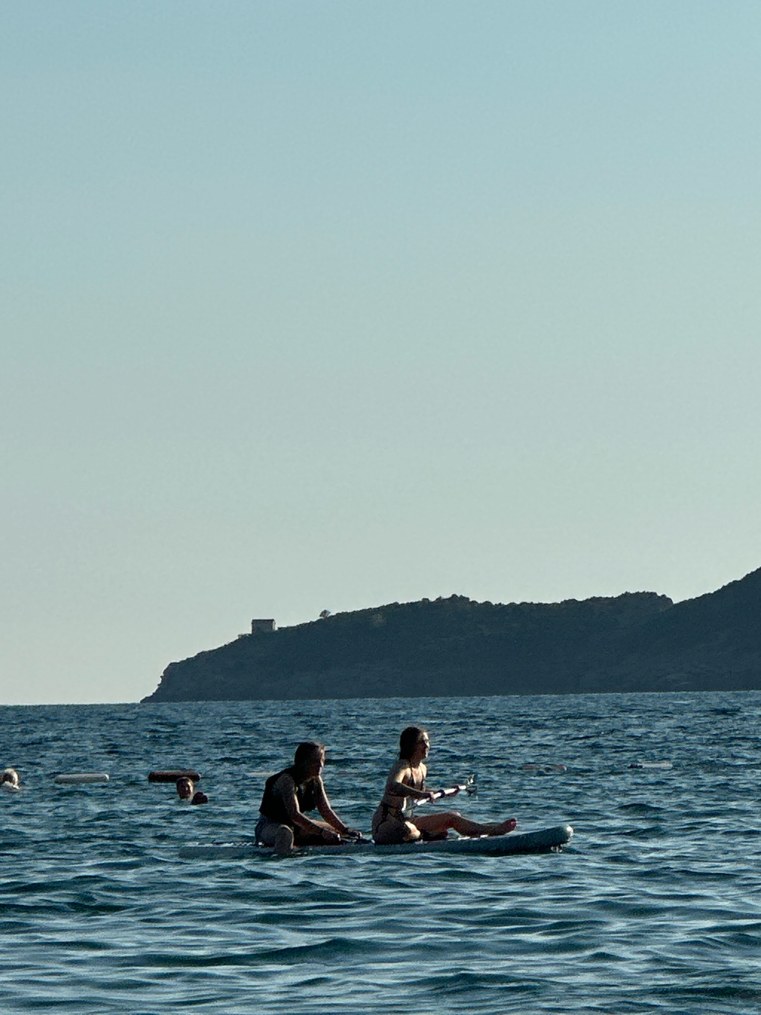 In the city of Boreti, Montenegro, two people are on a paddleboard in the water. The male, on the left, is wearing a dark vest and light shorts. He is looking to his right, towards the female on the paddleboard. The female, on the right, is wearing a patterned swimsuit and is holding a paddle. She is looking to her left. The water is choppy and blue. In the background, there is a dark hill with a building on top and a clear, pale blue sky. There are also other people in the water in the distance, some floating and some with their heads visible.
