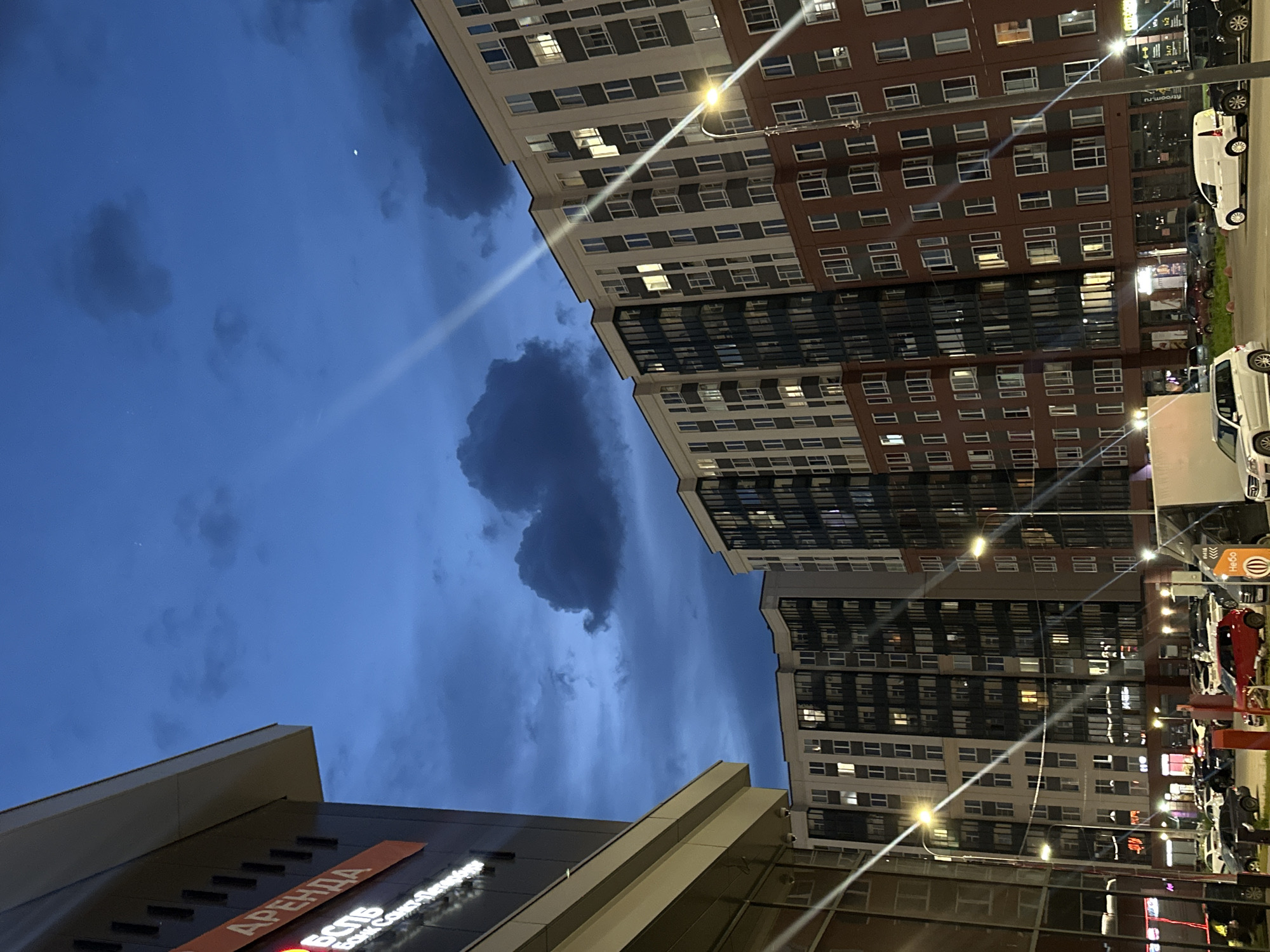 The image captures an urban environment at dusk or night in Murino, Russia. The sky is a deep blue, partially covered by dark, scattered clouds, with a faint crescent moon visible in the upper right quadrant. Multiple bright lens flares emanate from streetlights, cutting across the frame.
On the left, a commercial building with a dark facade and a light-colored overhang features an illuminated orange sign displaying the Cyrillic word "АРЕНДА" (RENT). Below it, in white Cyrillic text, are "ВСЛБ" and "Банк Санкт-Петербург" (Bank Saint Petersburg).
Across the center and right, multiple modern, multi-story residential buildings form a curved architectural line. These buildings feature facades in varying colors, including beige, dark grey, and dark reddish-brown, with numerous rectangular windows. A significant number of windows are illuminated, indicating interior lights are active.
At ground level, a parking area or street is occupied by numerous parked vehicles, including several white vans, a red car, and a silver SUV. One white van prominently displays the Cyrillic word "Небо" (Sky) on its side. Multiple streetlights with bright yellow-orange light sources are present. On the ground floor of the residential buildings, commercial establishments are indicated by illuminated signs, including "FITROOM.RU", "24/7", and "1+2". No human figures are discernible within the scene.