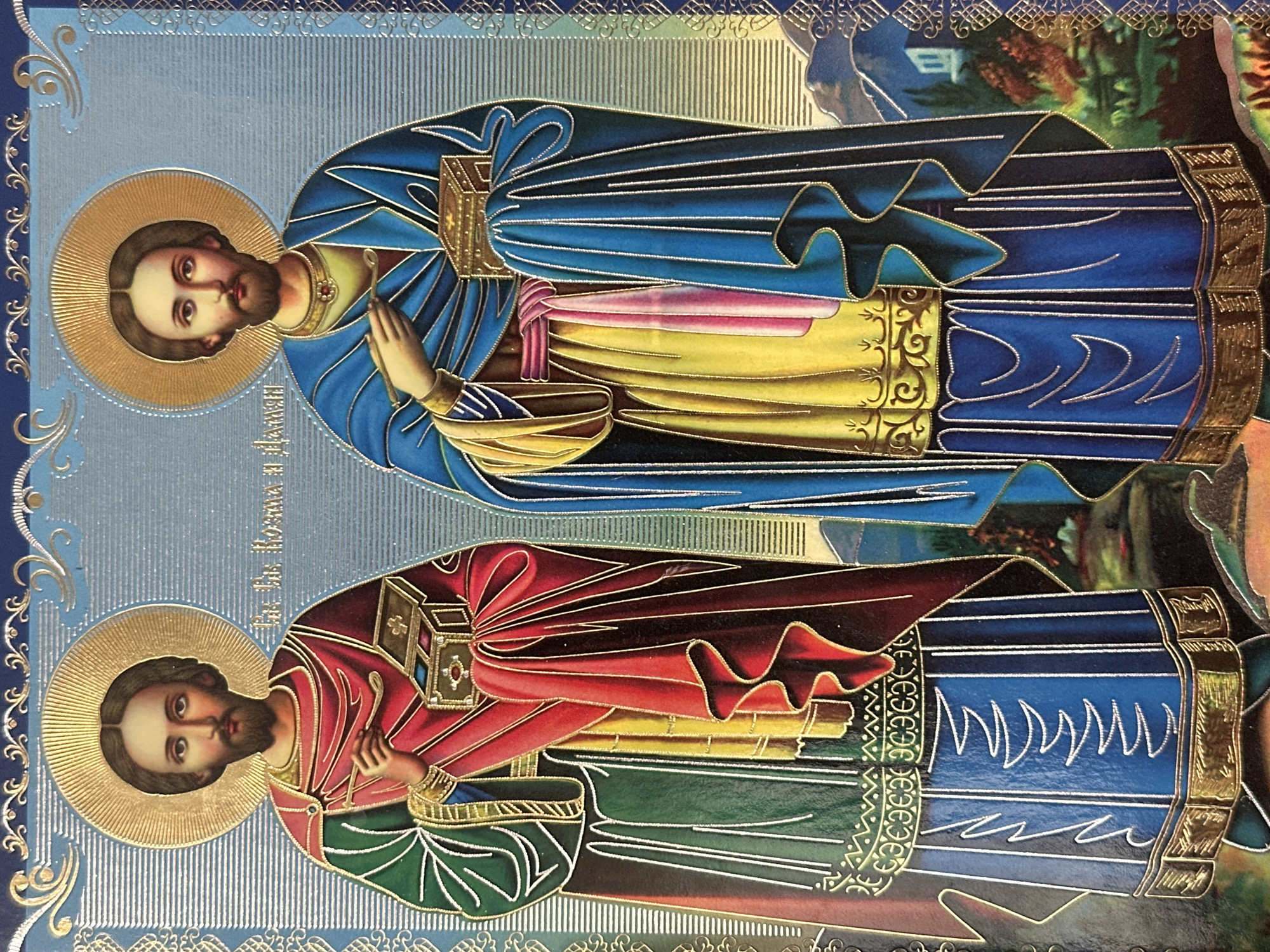 The image is an icon depicting two male figures, likely saints, dressed in richly colored robes. Both figures have halos and beards, and are holding ornate boxes. The figure on the left wears a green outer robe over a red and yellow garment, while the figure on the right wears a blue outer robe over a yellow garment with pink accents. The background features a landscape with mountains, trees, and a building, rendered in a stylized, traditional iconographic manner. The overall impression is one of religious art. There is no indication of the location in the city of Marten, Bulgaria.