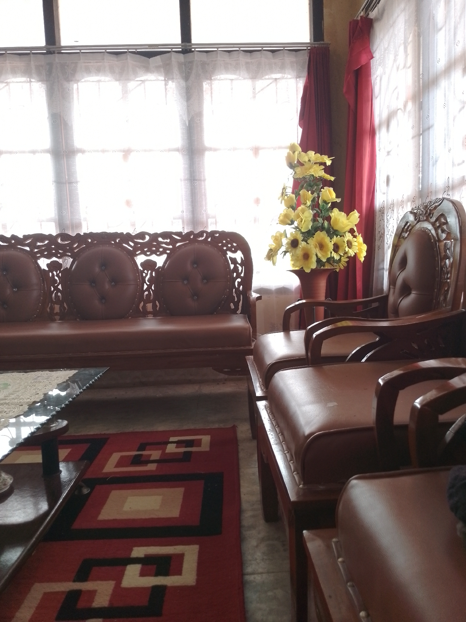 The image shows a living room with brown leather furniture, a patterned rug, and a vase of yellow flowers. The room is lit by natural light coming through a window with white curtains and red drapes. The furniture includes a sofa with carved wooden trim and three round tufted cushions, and two armchairs. A coffee table with a dark wood base and a textured surface is positioned in front of the sofa. The rug on the floor is red with a geometric pattern of white and black squares and rectangles. A large vase filled with artificial yellow flowers stands on the floor. The overall impression is one of a cozy and somewhat traditional interior.

This scene is likely located in a home in Bukittinggi, Indonesia. Bukittinggi is known for its traditional Minangkabau architecture and rich cultural heritage, and this living room's furnishings, particularly the carved wood elements, could be indicative of local craftsmanship. The use of leather furniture and patterned rugs is common in homes across Indonesia. The bright natural light suggests the image was taken during the daytime.