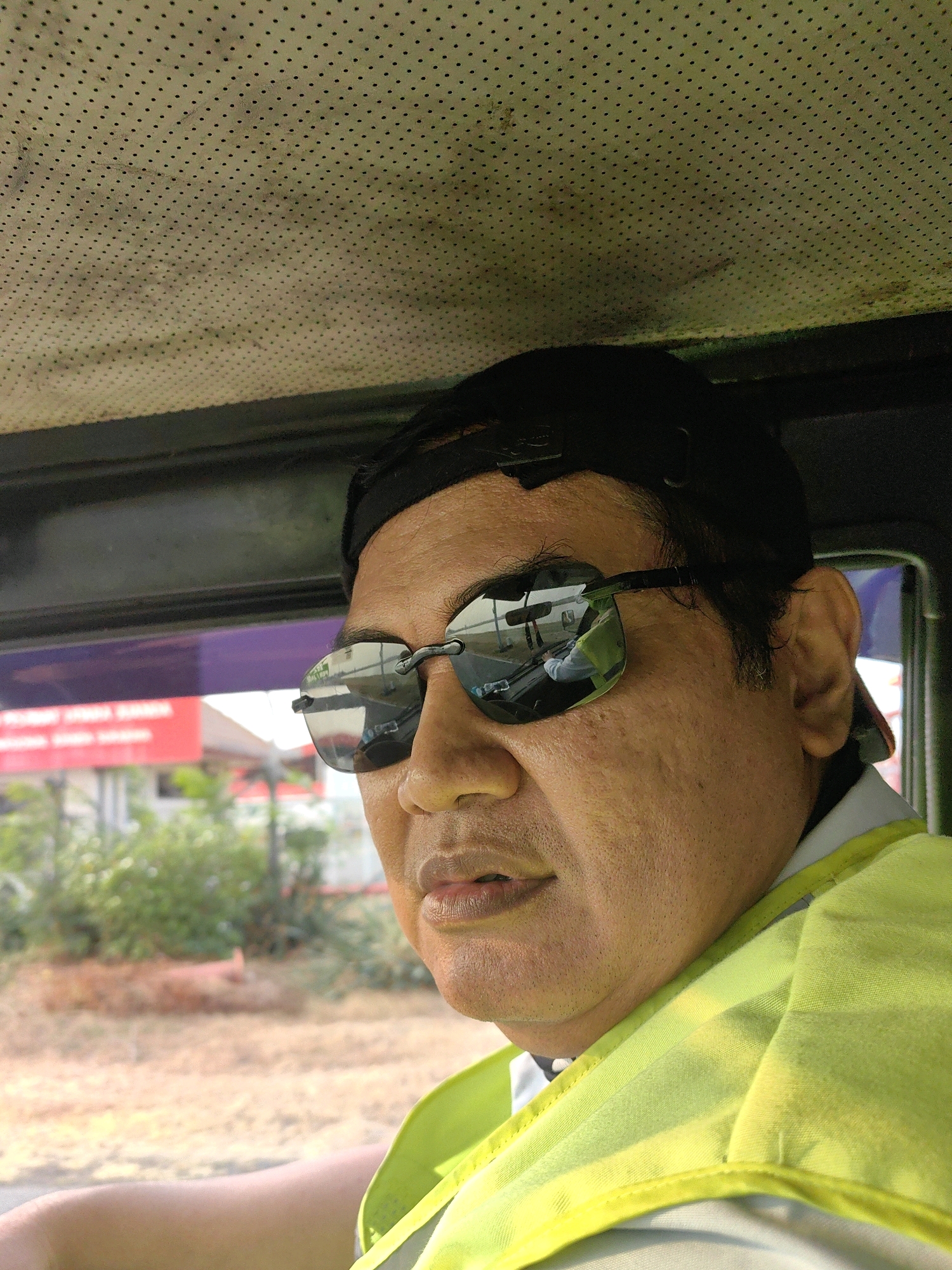 A man wearing sunglasses and a bright yellow vest is sitting in the passenger seat of a vehicle. The man has dark hair and is looking forward. The vehicle's interior is visible, with a perforated ceiling and dark trim. Through the window, a blurred outdoor scene can be seen, with trees and buildings in the background. The man is likely in Pranti, Indonesia, given the context provided.