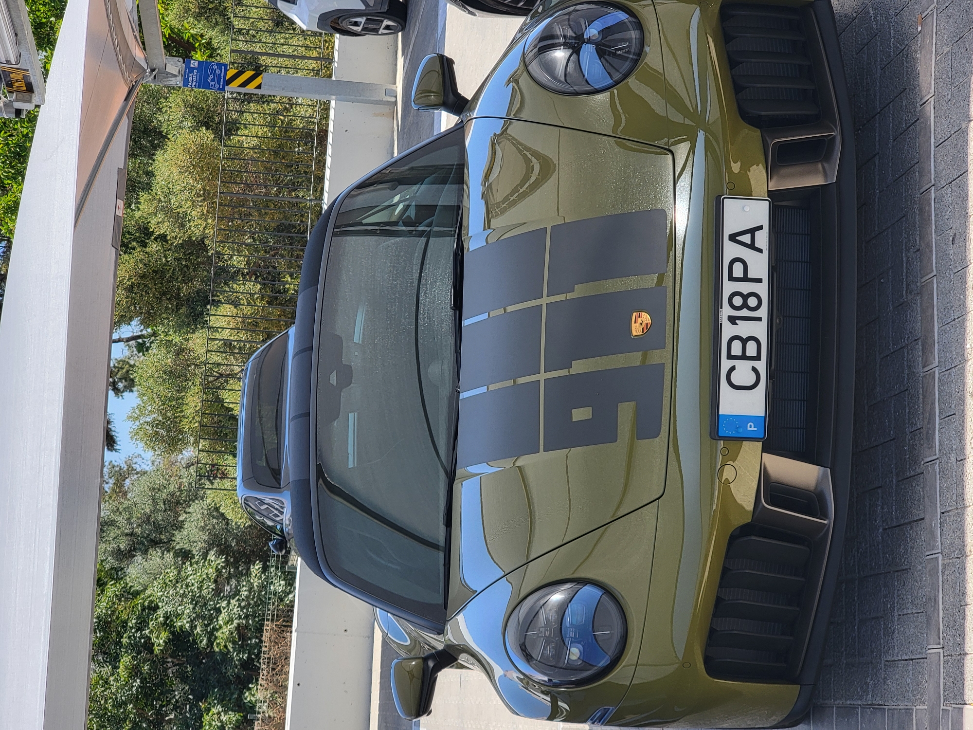 This is a frontal, slightly low-angle, medium shot of a green Porsche 911 convertible parked outdoors under a white awning. The car has a black soft top, a black racing stripe pattern with "911" emblazoned on the hood, and a Portuguese license plate reading "CB 18 PA". The car is positioned on a paved area with a cobblestone-like pattern. Behind the Porsche, another car, possibly also a Porsche, is visible, and further back, a white SUV is parked. In the background, there are trees and a metal fence. A blue and yellow sign is attached to a post to the right of the cars, indicating a private parking area. The scene is brightly lit, suggesting it is daytime. The location is identified as Quarteira, Portugal. There are no people visible in the image.