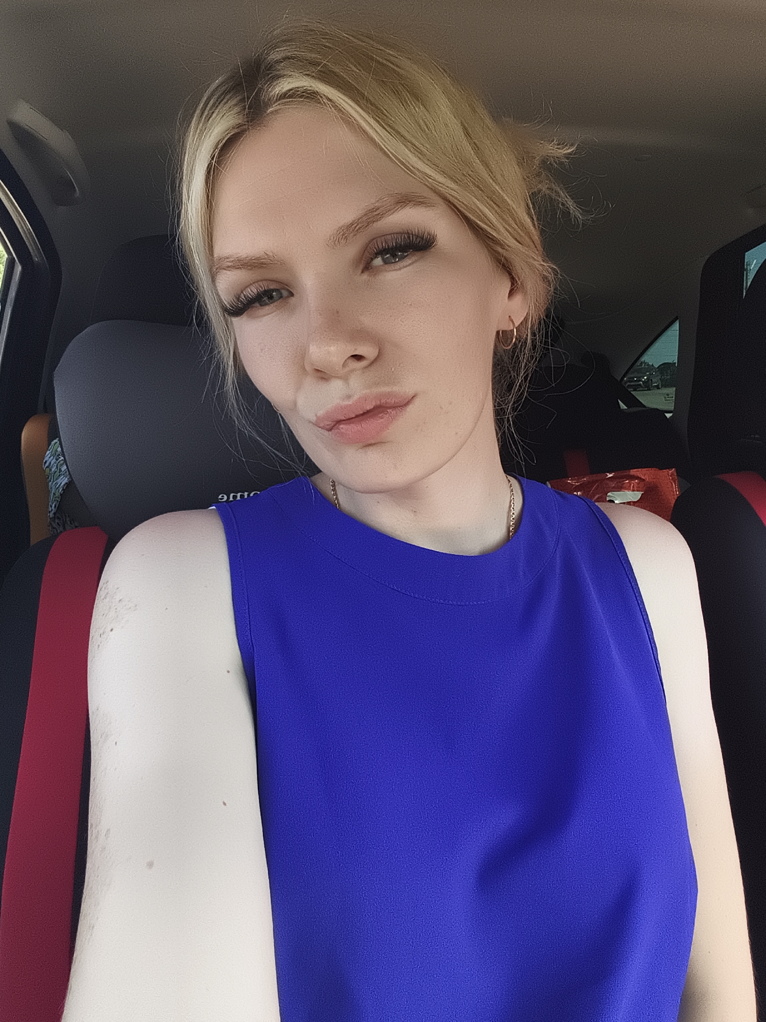 A young woman with blonde hair and fair skin is taking a selfie in a car. She is wearing a bright blue sleeveless top and a thin gold necklace. Her left arm is resting on the car seat, and her right shoulder is visible. The background shows the interior of the car, including dark seats and a glimpse of the outdoors through the windows. The image appears to have been taken during the day. There is no discernible information to suggest the location is in the city of Ягул, Russia.