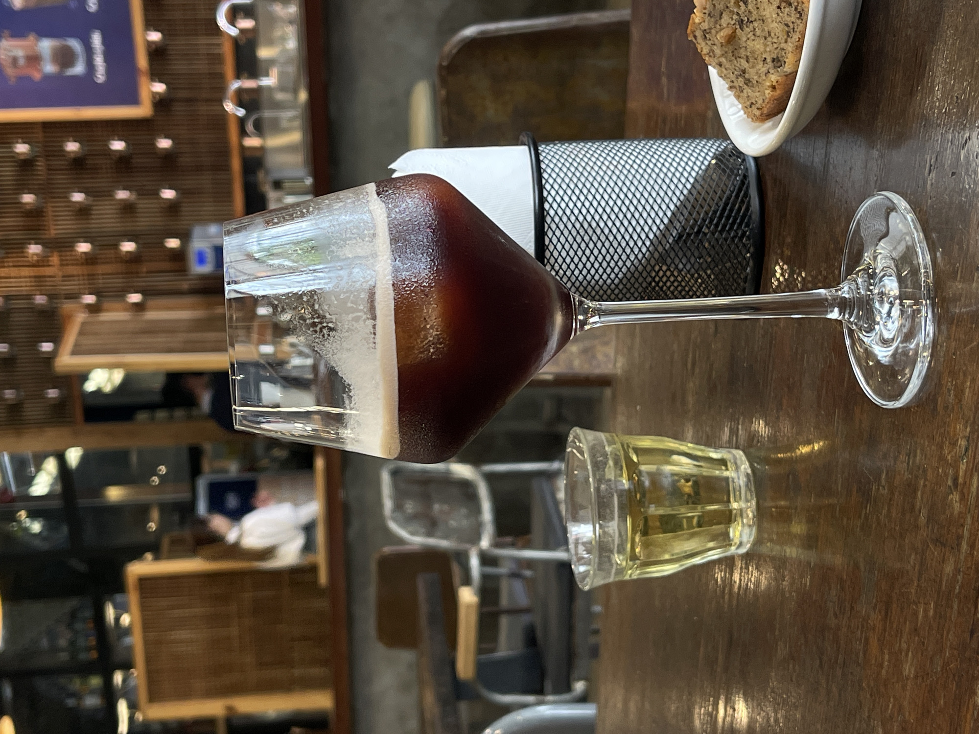 The scene is an indoor setting, likely a café in Da Lat, Vietnam. A stemmed glass of dark liquid with a creamy top sits on a dark wooden table. A small glass of amber liquid is next to it. Beside the glass is a black wire napkin holder with white napkins. A partially eaten piece of banana bread is in a white dish. The background features a partially visible menu board and several blurred tables and chairs with a person in a white shirt. The lighting is soft and diffused.