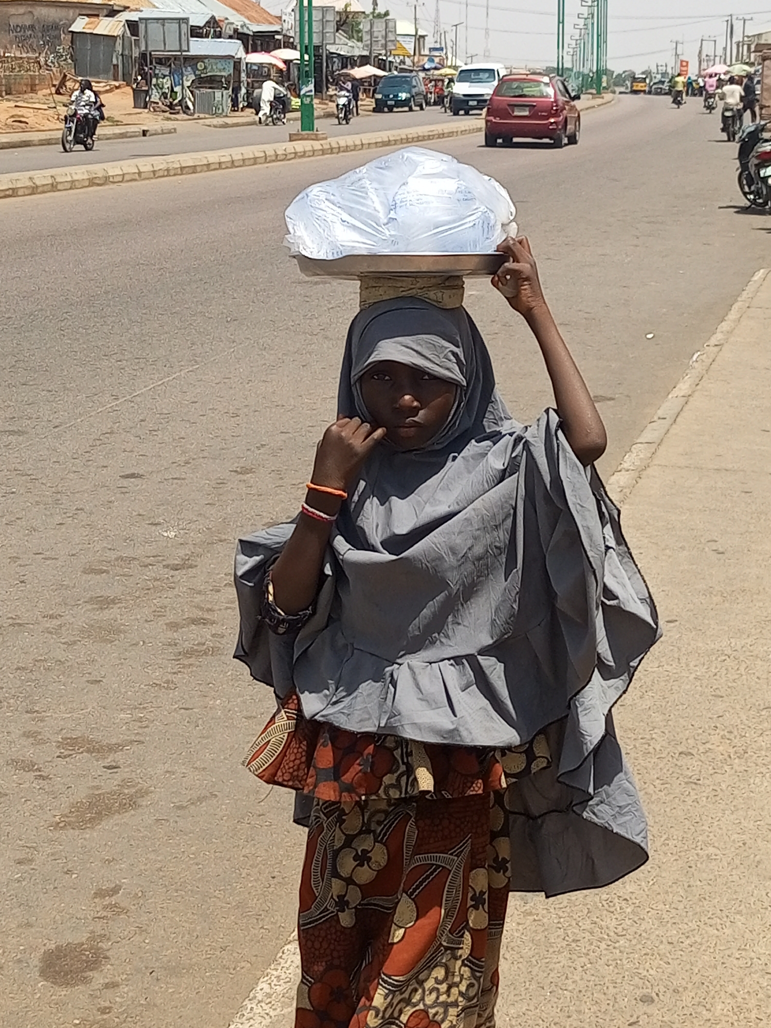 A young girl, likely a vendor, walks on the side of a paved road in Gombe, Nigeria. She wears a grey hijab and a colorful patterned skirt. On her head, she balances a large, clear plastic bag filled with what appears to be water bottles, resting on a metal tray. Her right arm is slightly raised, fingers bent as if steadying the load. Traffic, including cars and motorcycles, moves on the road behind her. Buildings and market stalls line the left side of the street, suggesting a commercial area. The sun casts distinct shadows, indicating a bright, daytime scene.